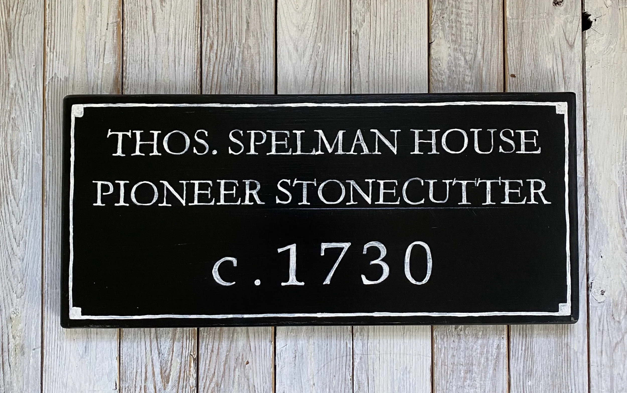 A black sign with white painted text reading: "THOS. SPELMAN HOUSE PIONEER STONECUTTER c. 1730" mounted on a weathered wooden wall.