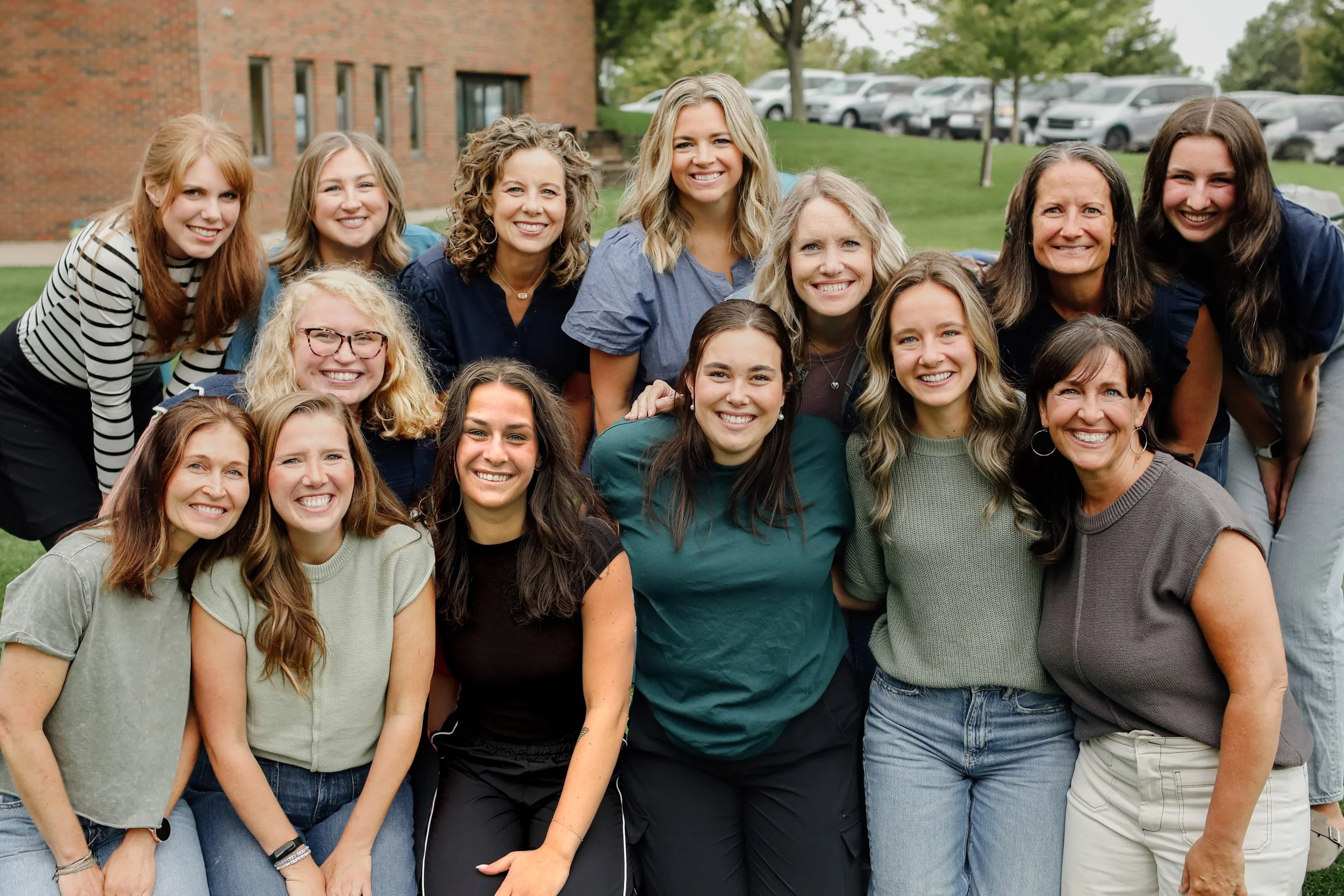 Group of women smiling outdoors in front of a green lawn and parking lot.