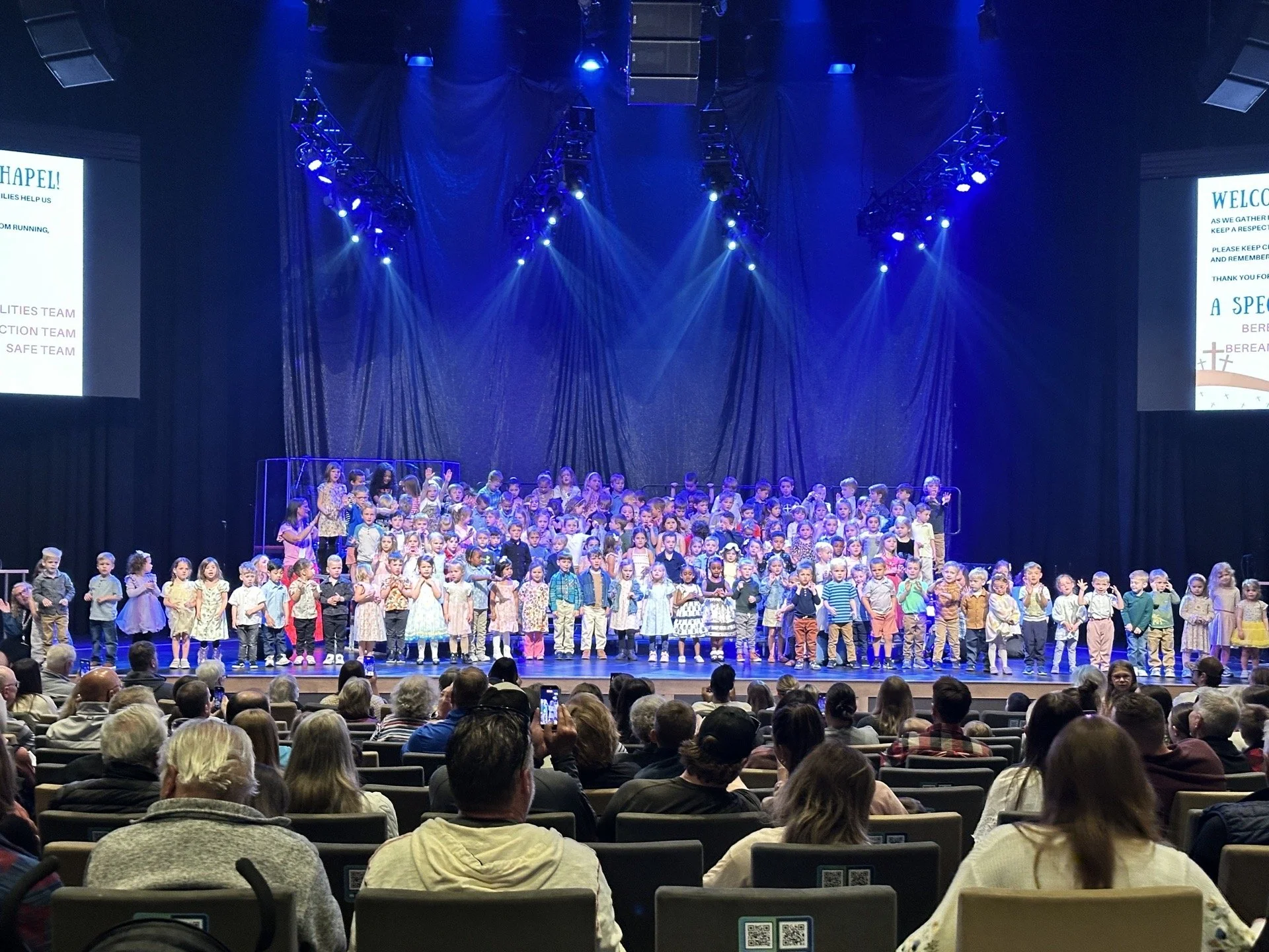 Children on stage performing or singing in front of an audience in a theater or auditorium, with two large screens displaying welcoming messages on either side of the stage and blue stage lights overhead.