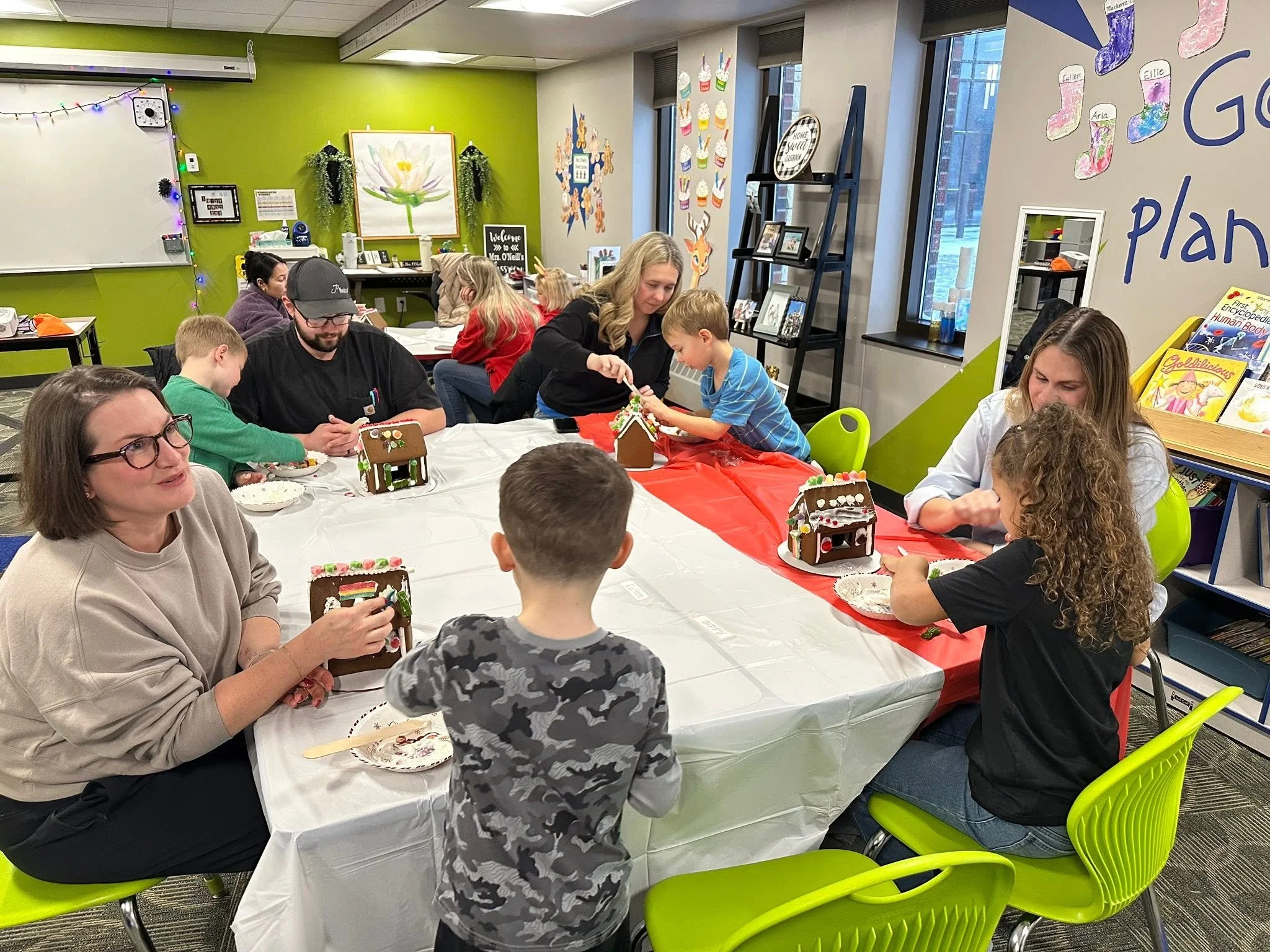 Children and adults gathered around a table decorated with a red tablecloth, making gingerbread houses. The room has bright walls decorated with festive and colorful holiday artwork, including cupcakes and Christmas stockings, indicating a holiday celebration.