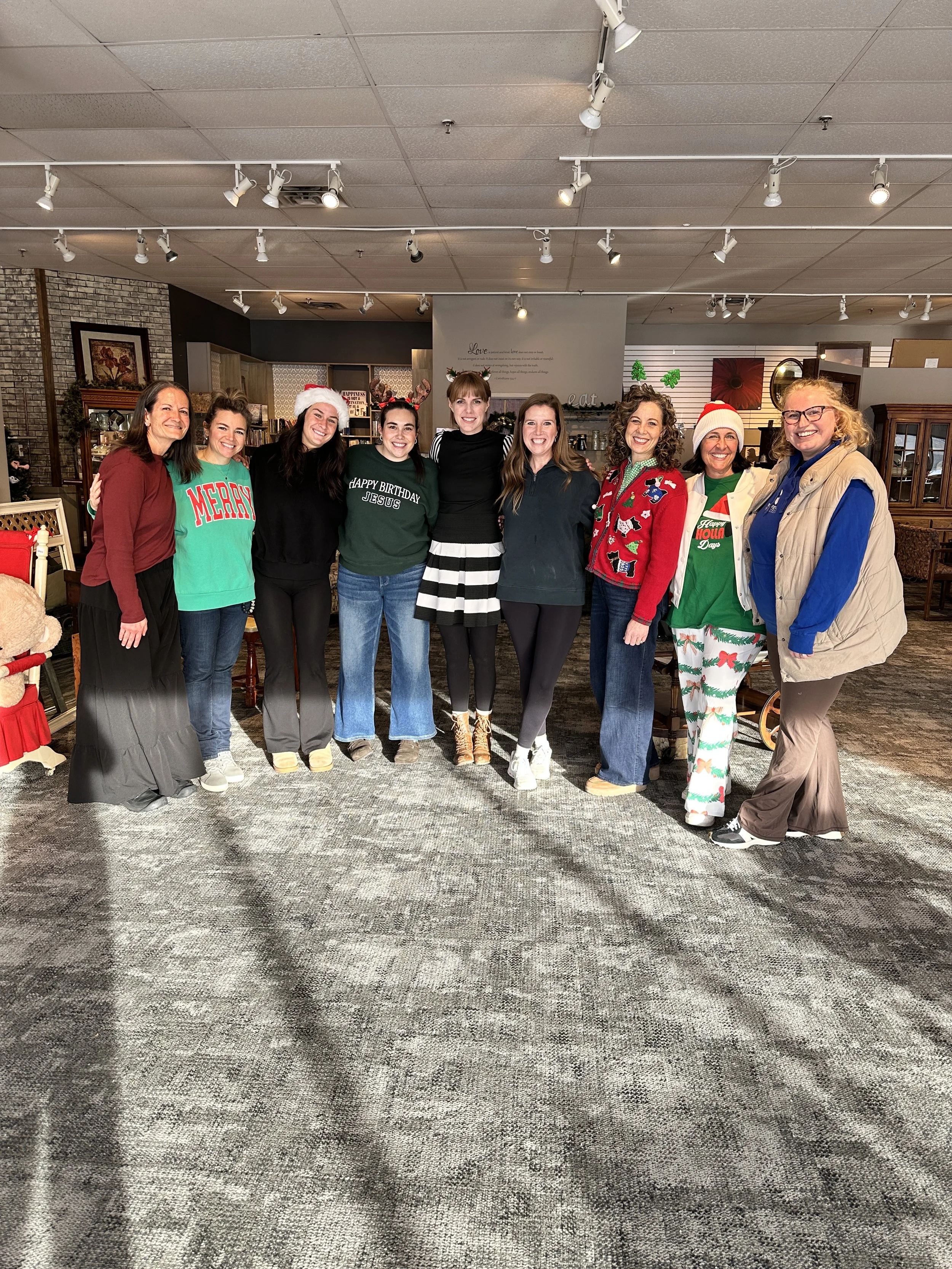 A group of nine women standing together indoors, dressed in Christmas-themed clothing and accessories, smiling for a group photo.