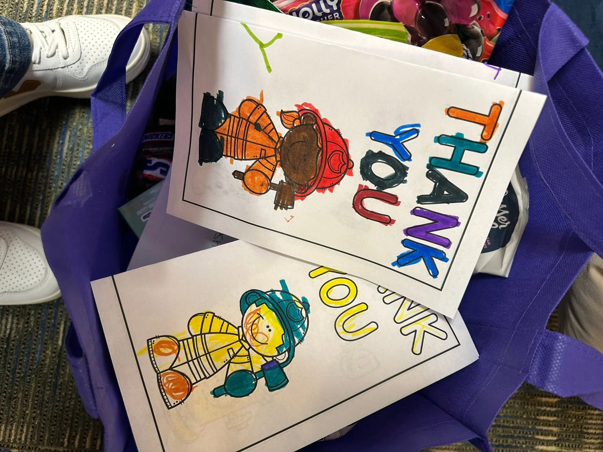 Coloring pages with a Thanksgiving theme, including a pilgrim girl and a turkey, placed inside a purple tote bag. In the background, a person wearing white sneakers and jeans is visible.