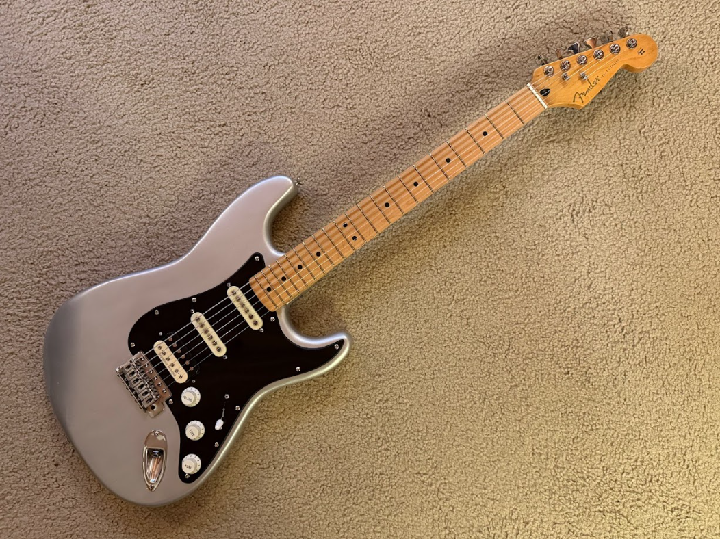 A silver electric guitar with a black pickguard, three single-coil pickups, and a maple neck, lying on a beige carpeted floor.