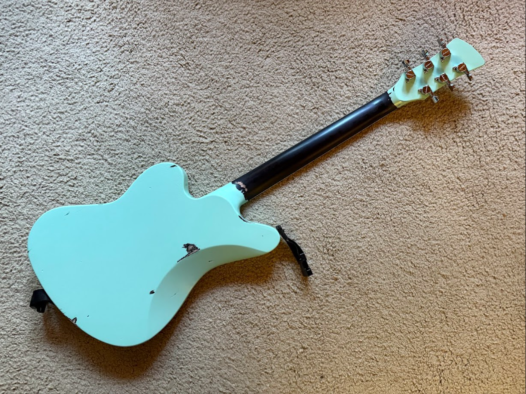 An electric guitar with a light blue body, black neck, and gold tuning pegs, lying on a beige carpet.