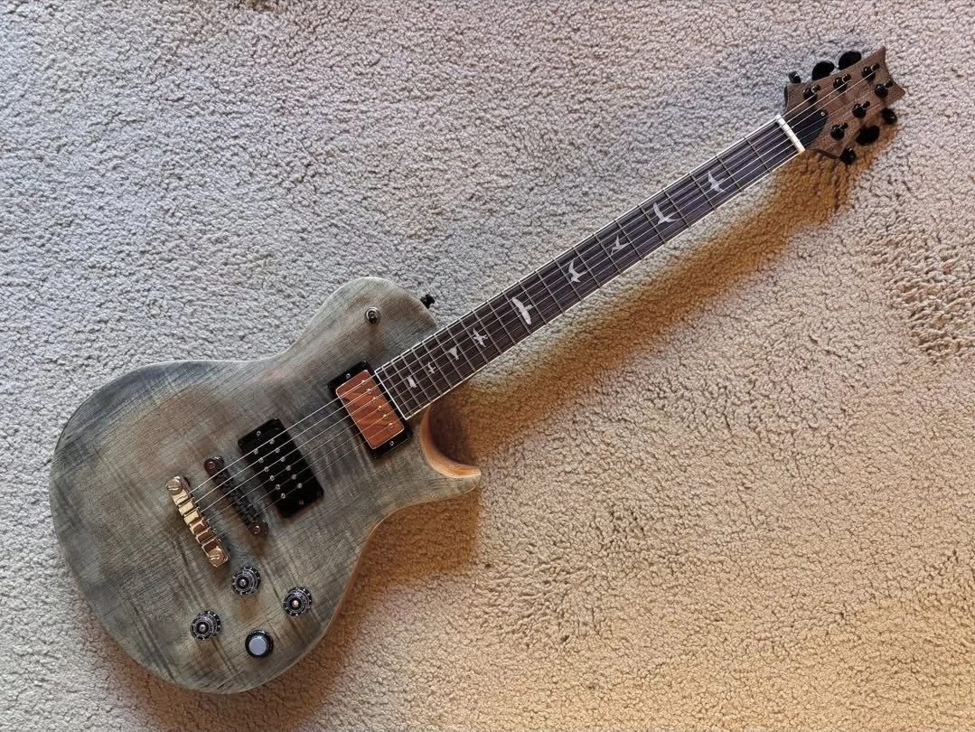 A grayish wooden electric guitar with a single-cutaway body shape and black hardware, lying on a beige carpeted floor.