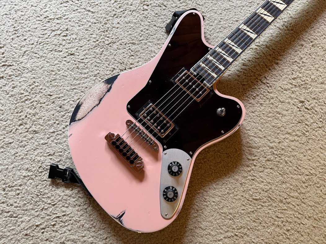 A pink and black electric guitar with a distressed finish, resting on a beige carpet.