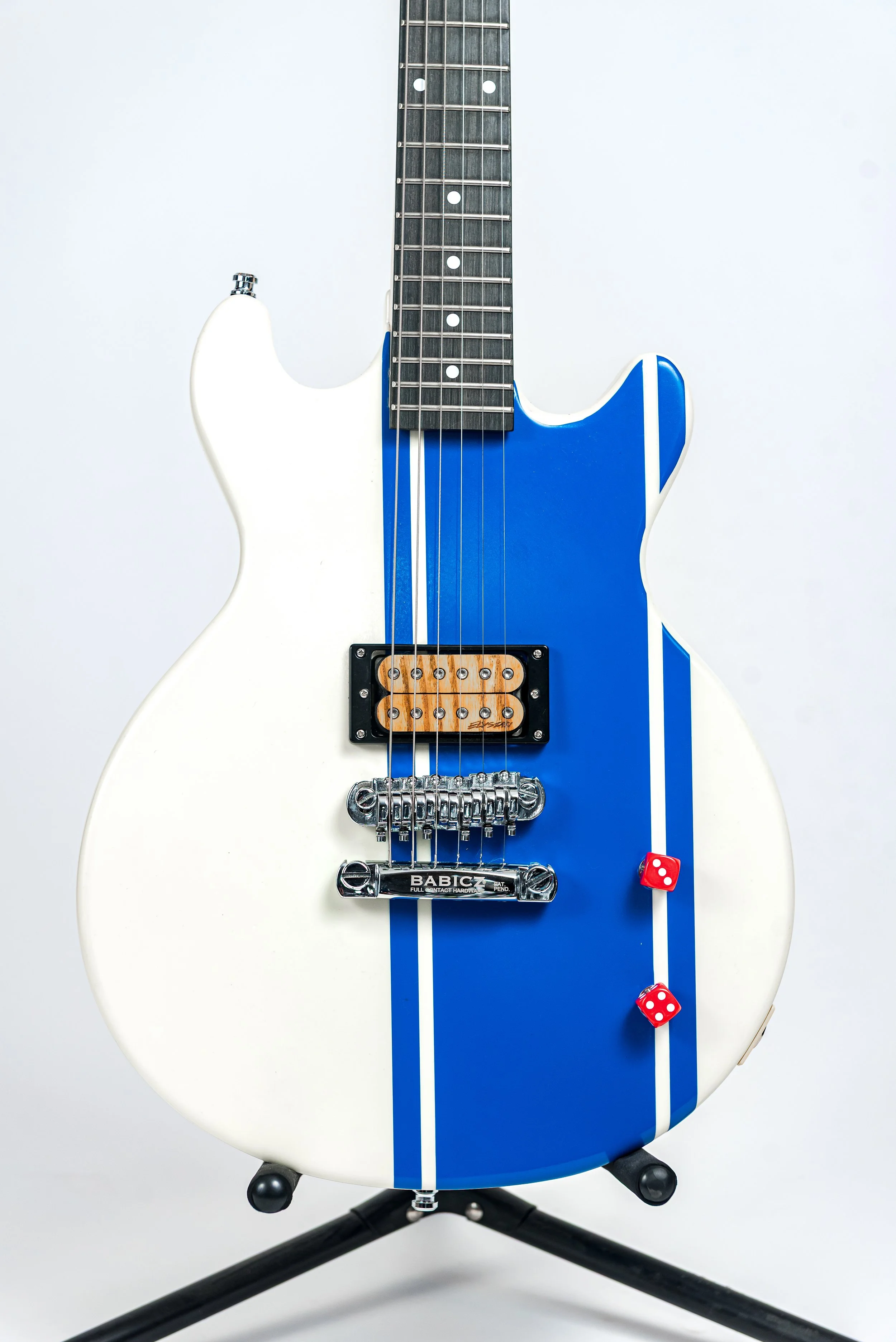 Electric guitar with a white body, blue pickguard, and two red dice attached, standing on a black tripod stand.