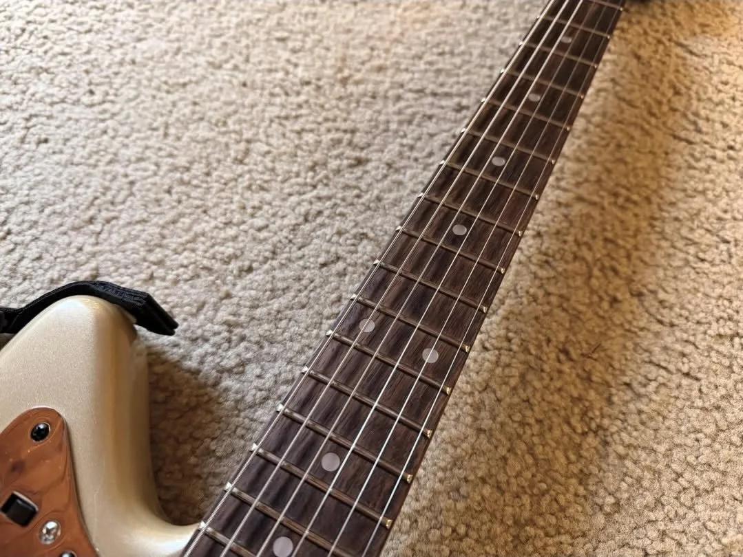 Close-up of an electric guitar neck with fretboard, strings, and dot inlays on the frets, resting on a carpeted floor.