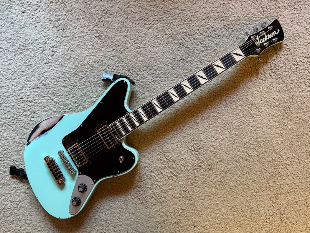 Blue electric guitar with a black pickguard, placed on a beige carpet, labeled 'Jackson' on the headstock.