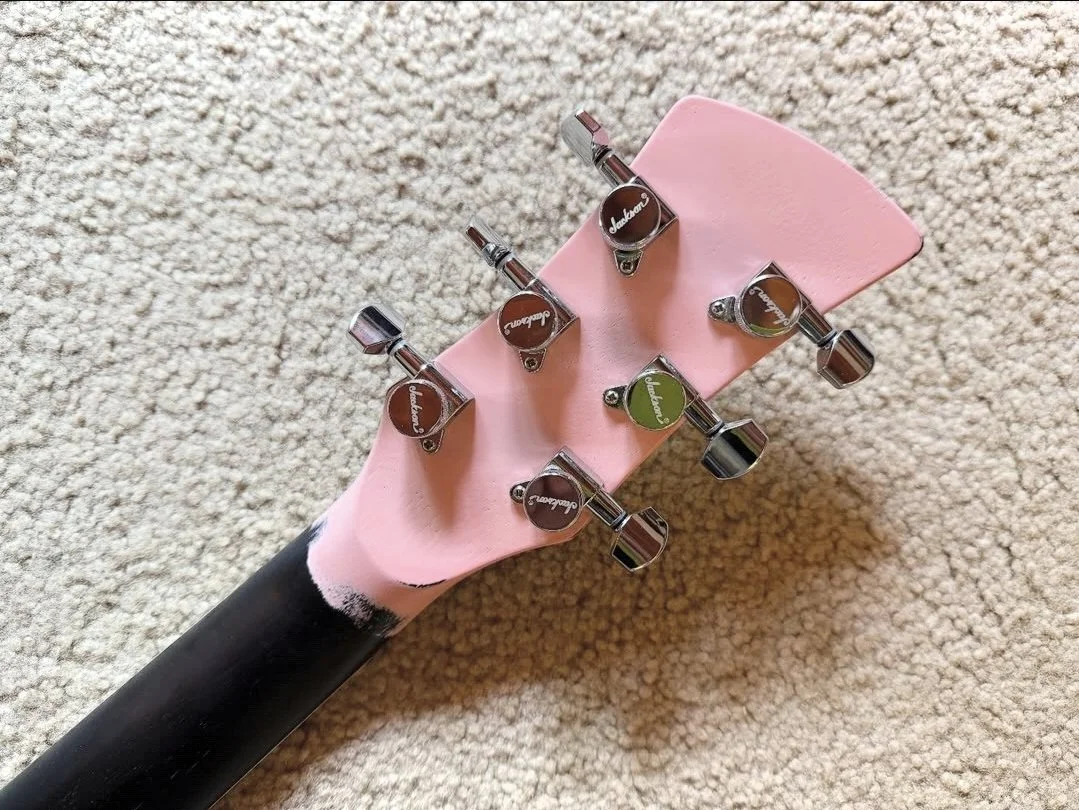 A black guitar neck with tuning pegs, placed on a beige carpet, with a pink foam piece covering the headstock.
