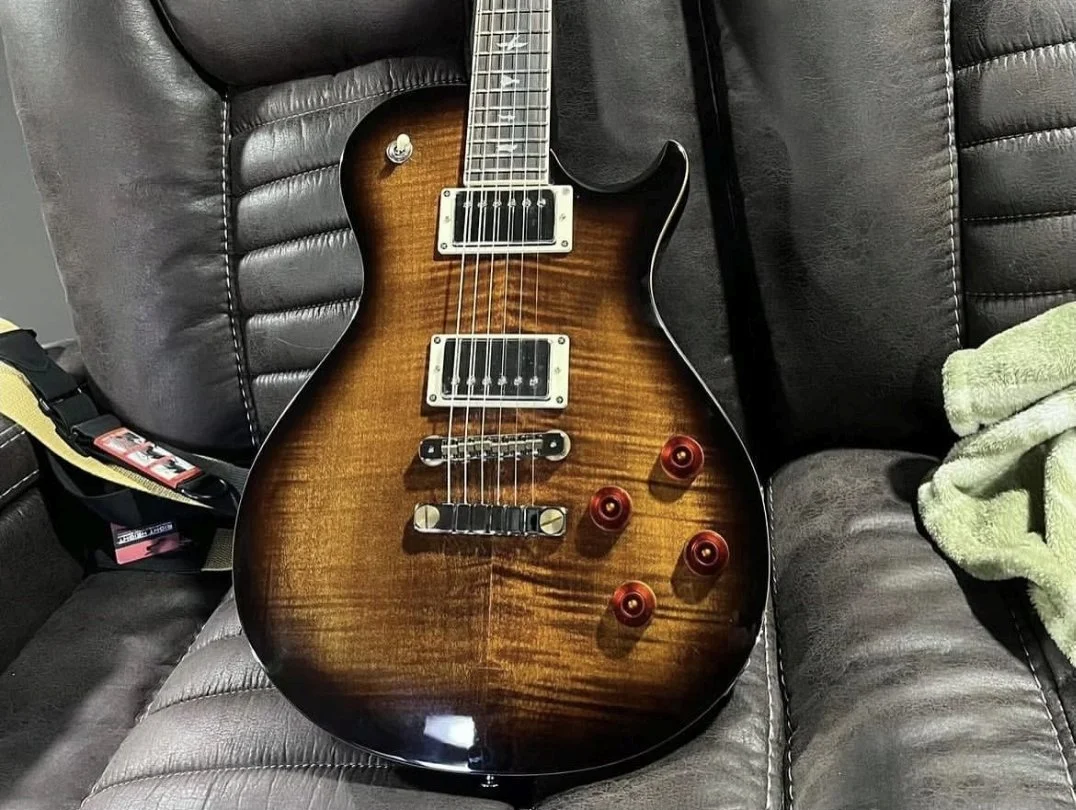 A sunburst electric guitar with a flamed wood top, silver hardware, and four control knobs resting on a dark leather couch.