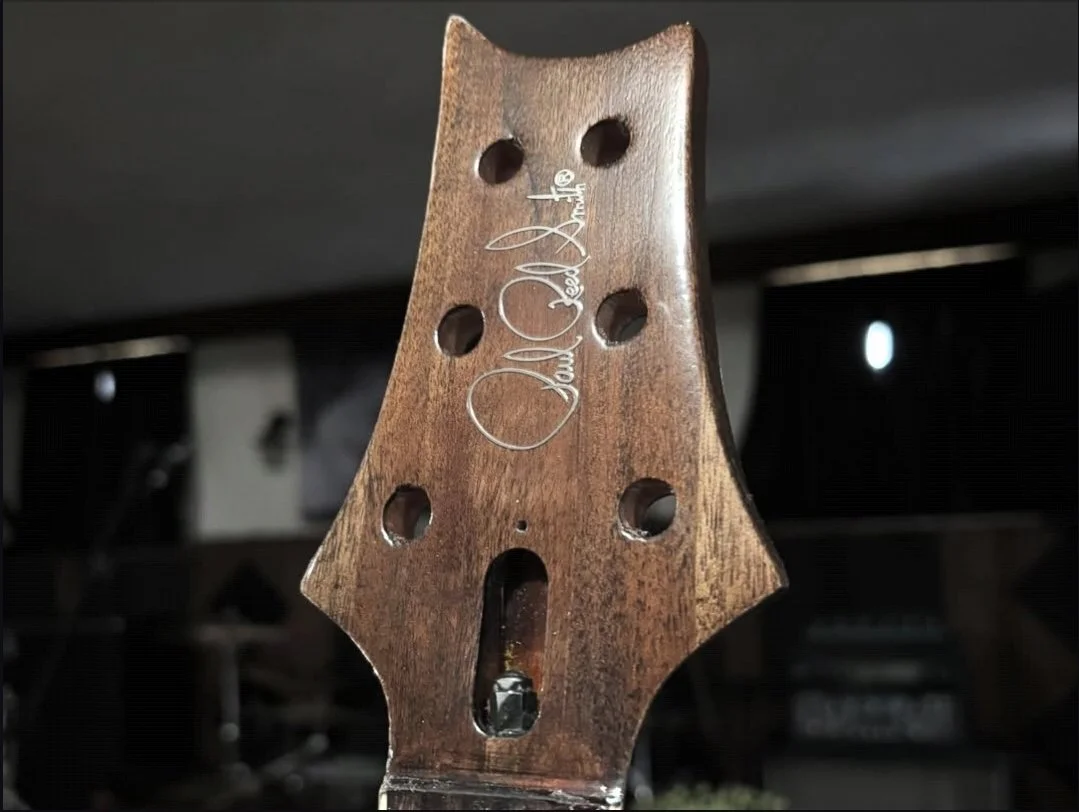 Close-up of a guitar headstock made of dark wood with a signature engraved on it.