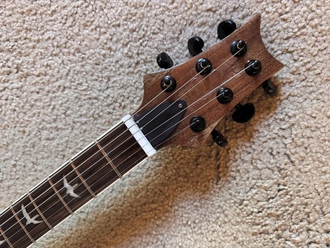 Close-up of a guitar headstock with black tuning pegs, a black pickguard, and custom inlays on the fretboard, resting on a beige carpet.