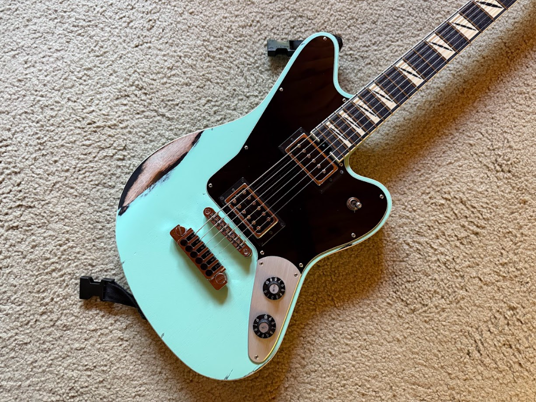 Electric guitar with a light blue body, black pickguard, and brown hardware, placed on a beige carpet.