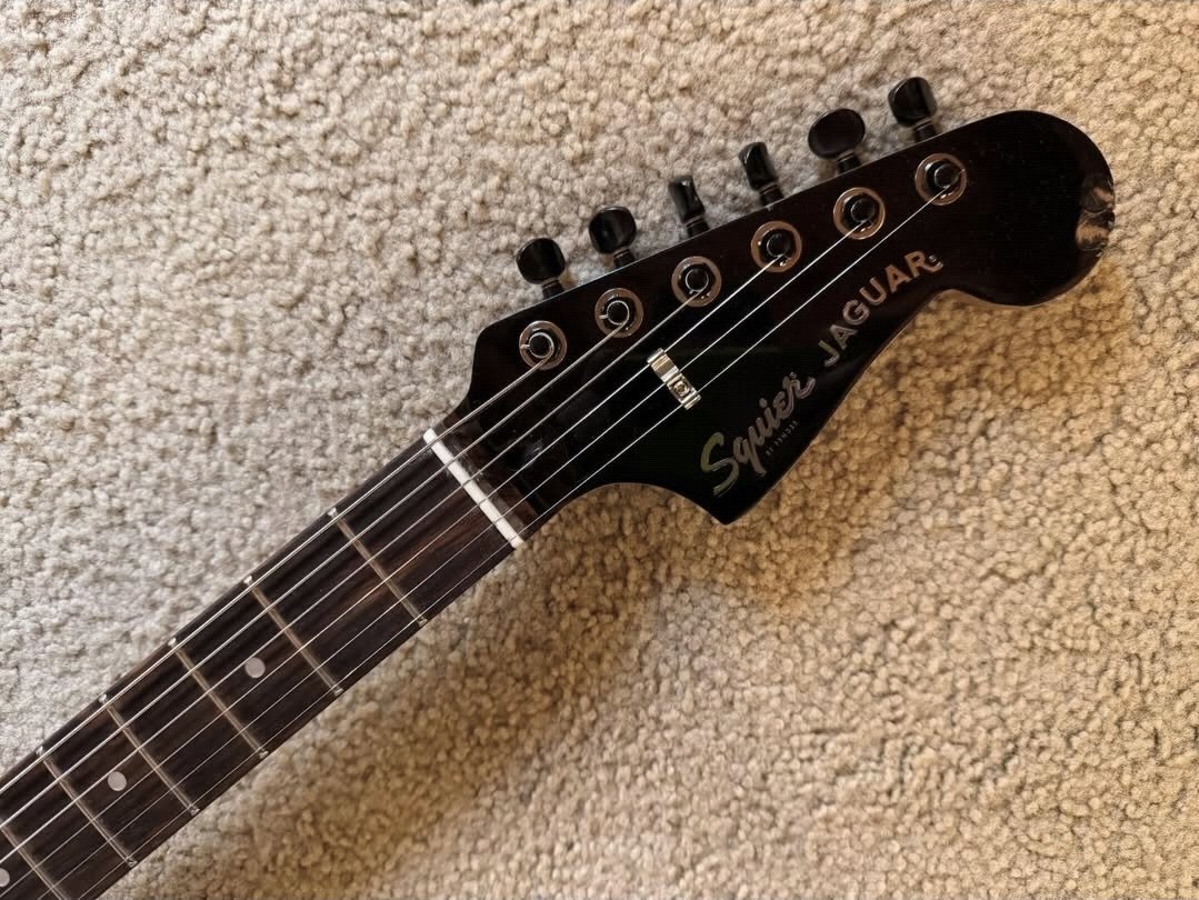 Close-up of a black Squier Jaguar electric guitar headstock with tuning pegs, mounted on a beige carpet.