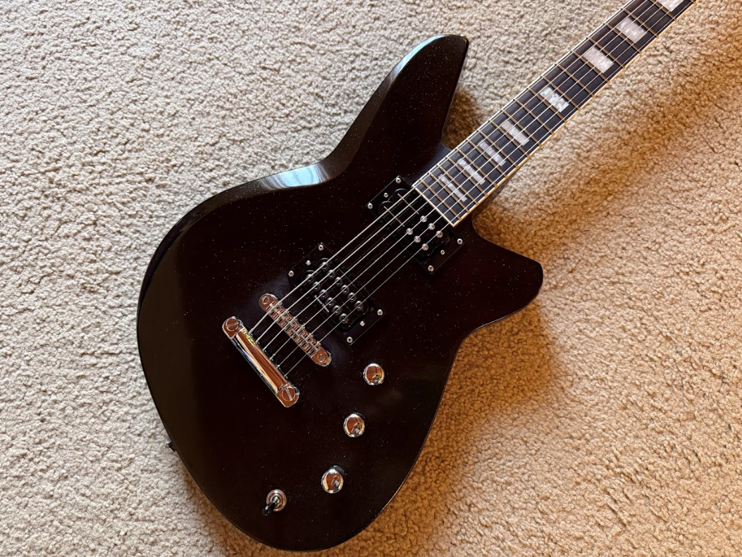 Black electric guitar with a sleek, modern design resting on a beige carpet.