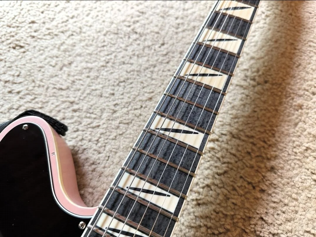 Close-up of an electric guitar neck and headstock on a beige carpet. The guitar has a black and pink body, with a distinctive fretboard featuring a decorative inlay pattern.
