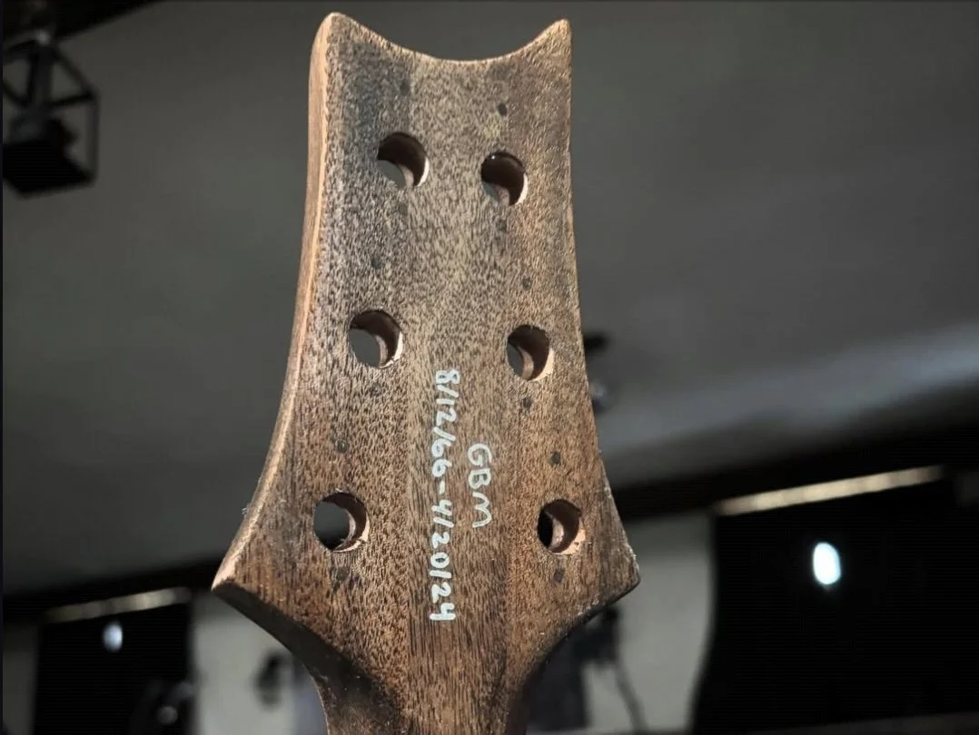 Close-up of a guitar headstock with six tuning peg holes, showing handwritten notes with date, measurements, and serial number.