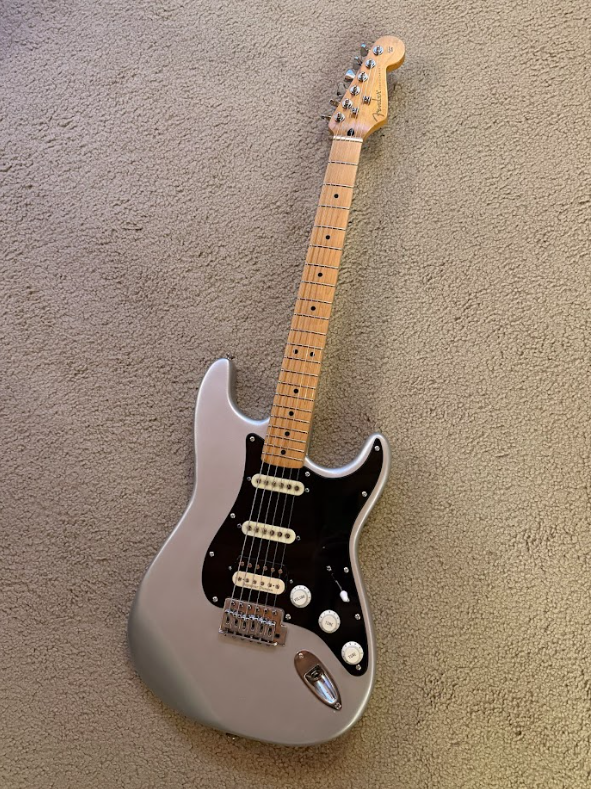 Silver electric guitar with a black pickguard, three pickups, a tremolo bridge, and a maple neck with a light finish, resting on a beige carpeted floor.