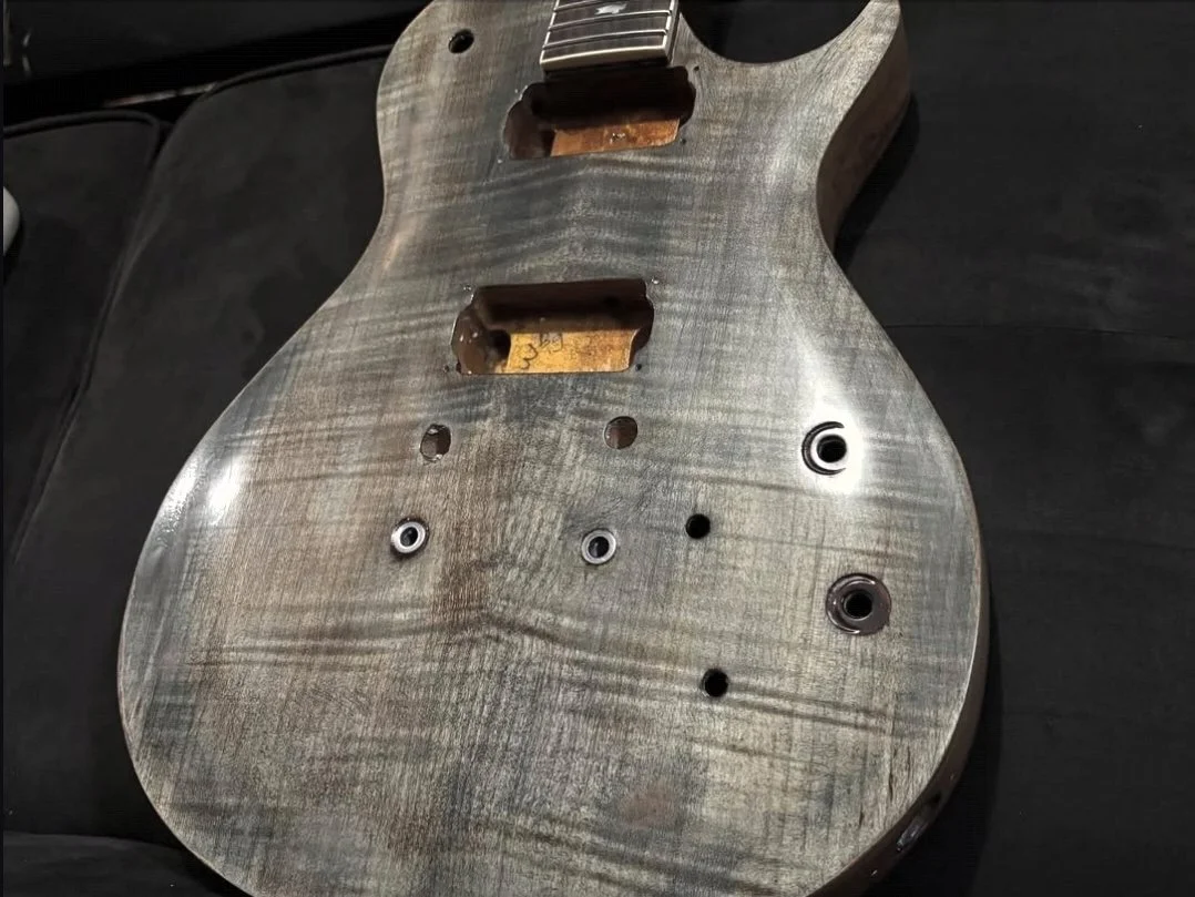 A wooden guitar body with no strings, showing the hollowed-out pickups and control cavities, resting on a black surface.