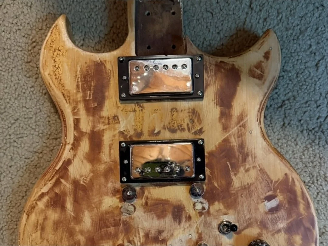 Close-up of a partially assembled electric guitar with a distressed wood finish, featuring two pickups, control knobs, and a toggle switch on a carpeted surface.
