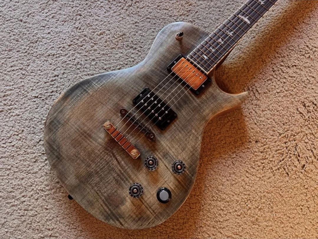 A wooden electric guitar with a natural wood finish, black pickups, and a set of control knobs, lying on a beige carpet.