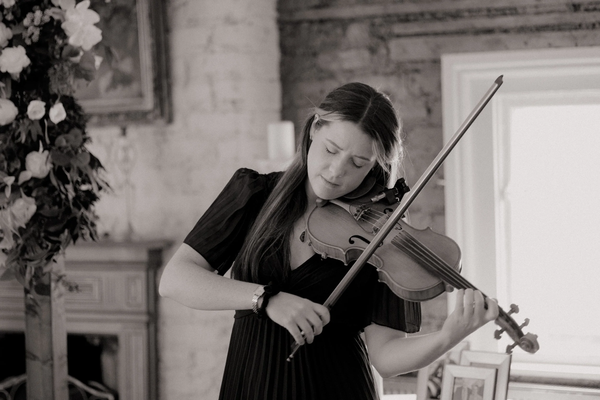 A woman playing the violin indoors, with a large bouquet of wedding flowers visible on the left and a window on the right in the background.