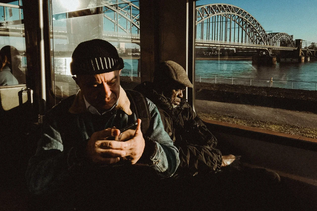 Two men sitting on a public transportation vehicle, with a bridge and water visible through the window behind them. One man, wearing a black beanie and denim jacket, is looking at his phone. The other man, wearing a flat cap and dark jacket, is resting with eyes closed.