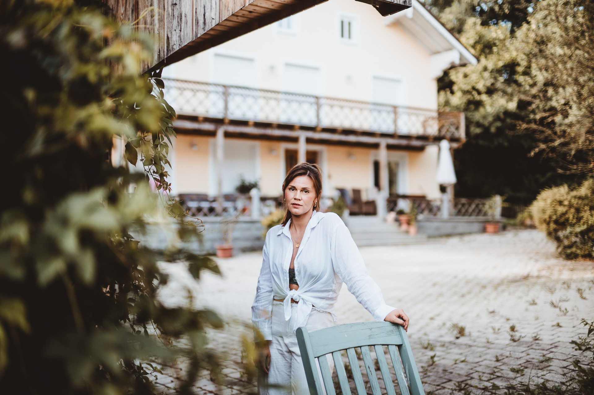 A woman standing outdoors on a patio with a house in the background, leaning on a wooden chair, wearing a white shirt tied at the waist and looking at the camera.