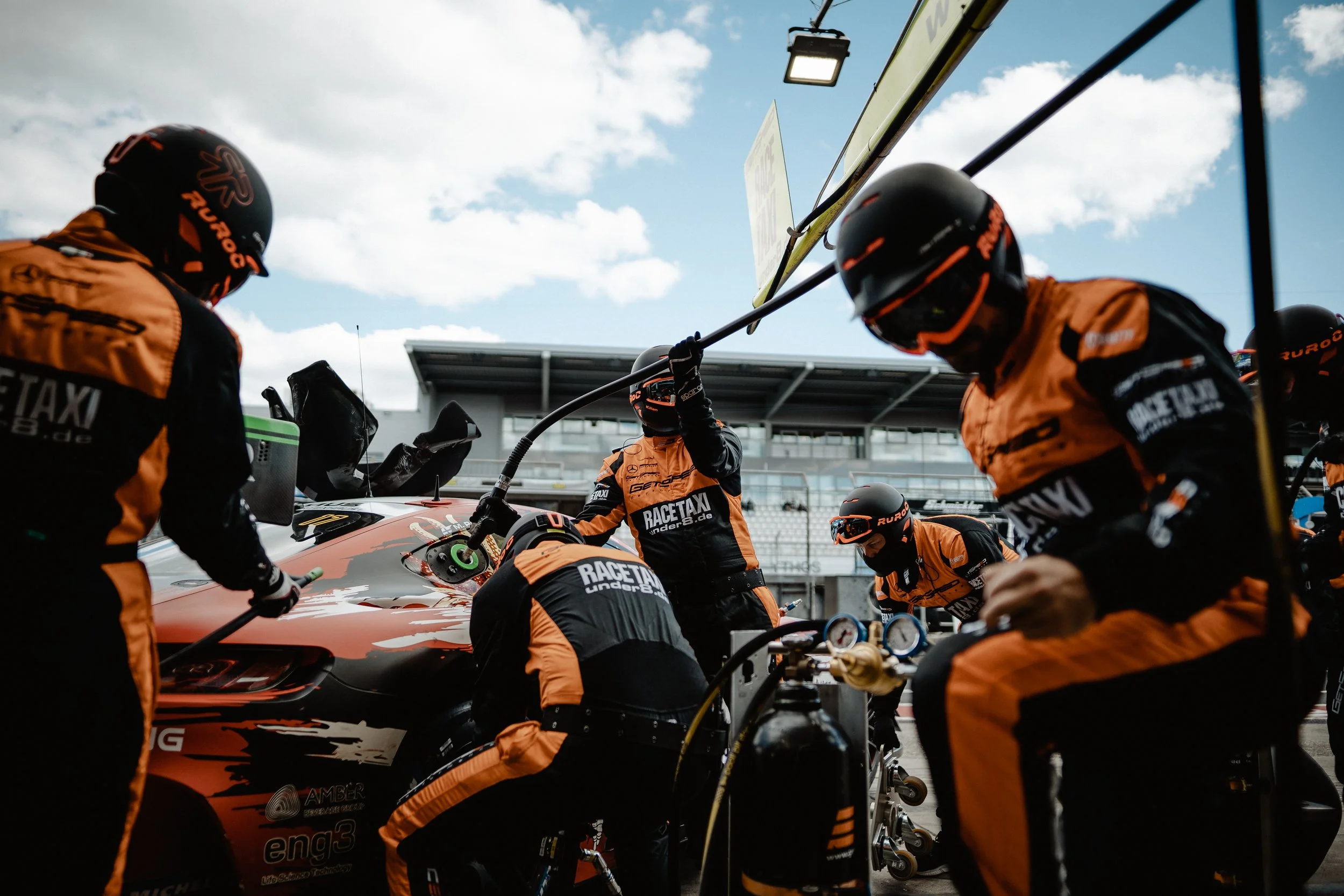 Race car team members in orange and black suits and helmets working on a race car in the pit lane, with a grandstand and cloudy sky in the background.