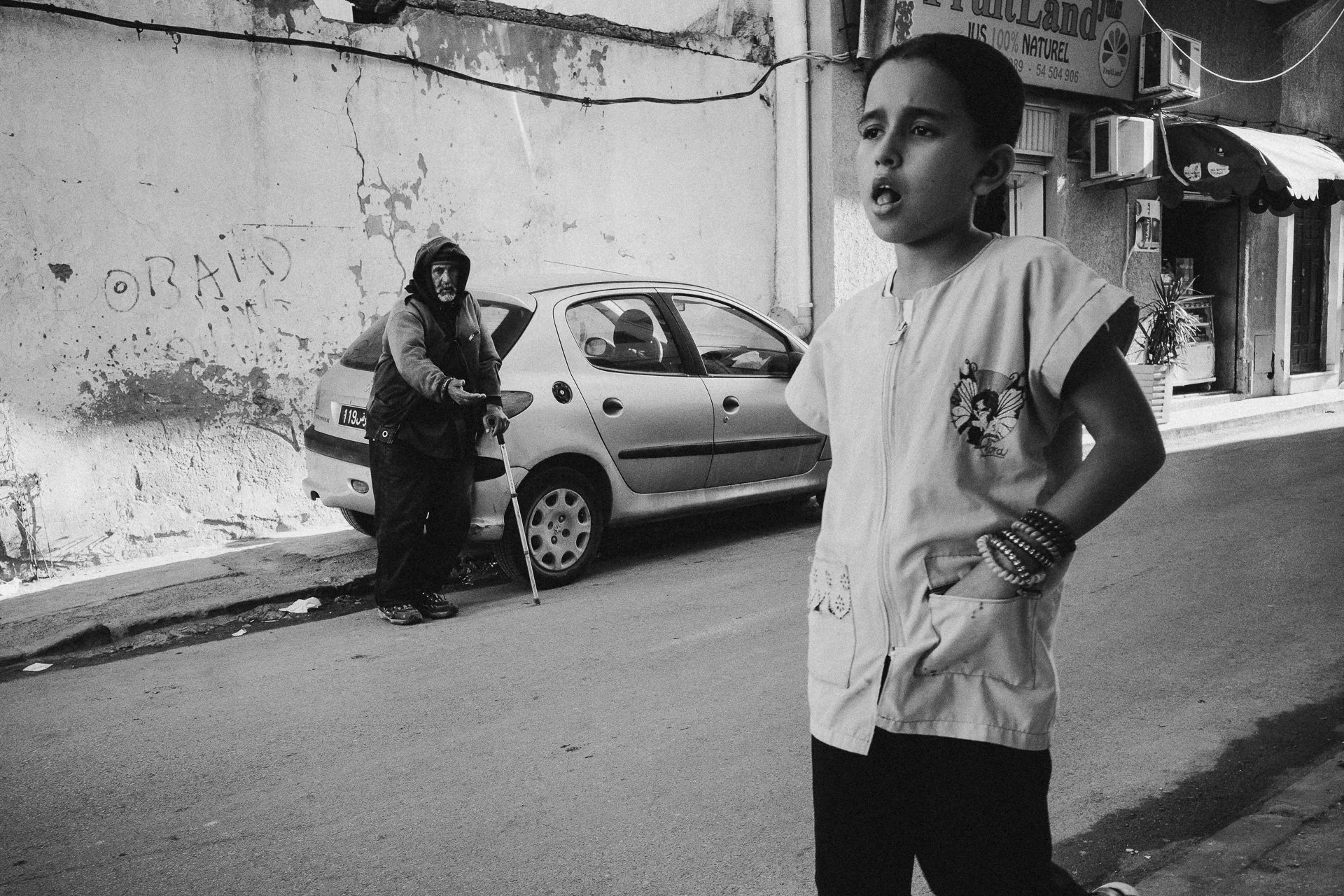 A young boy wearing a light-colored shirt and multiple bracelets stands on a street corner, looking towards the right with a curious expression. Behind him, an older woman with a mask and a hooded jacket leans on a cane beside a parked car, facing the camera. The background features a worn wall with graffiti and a storefront with air conditioning units.