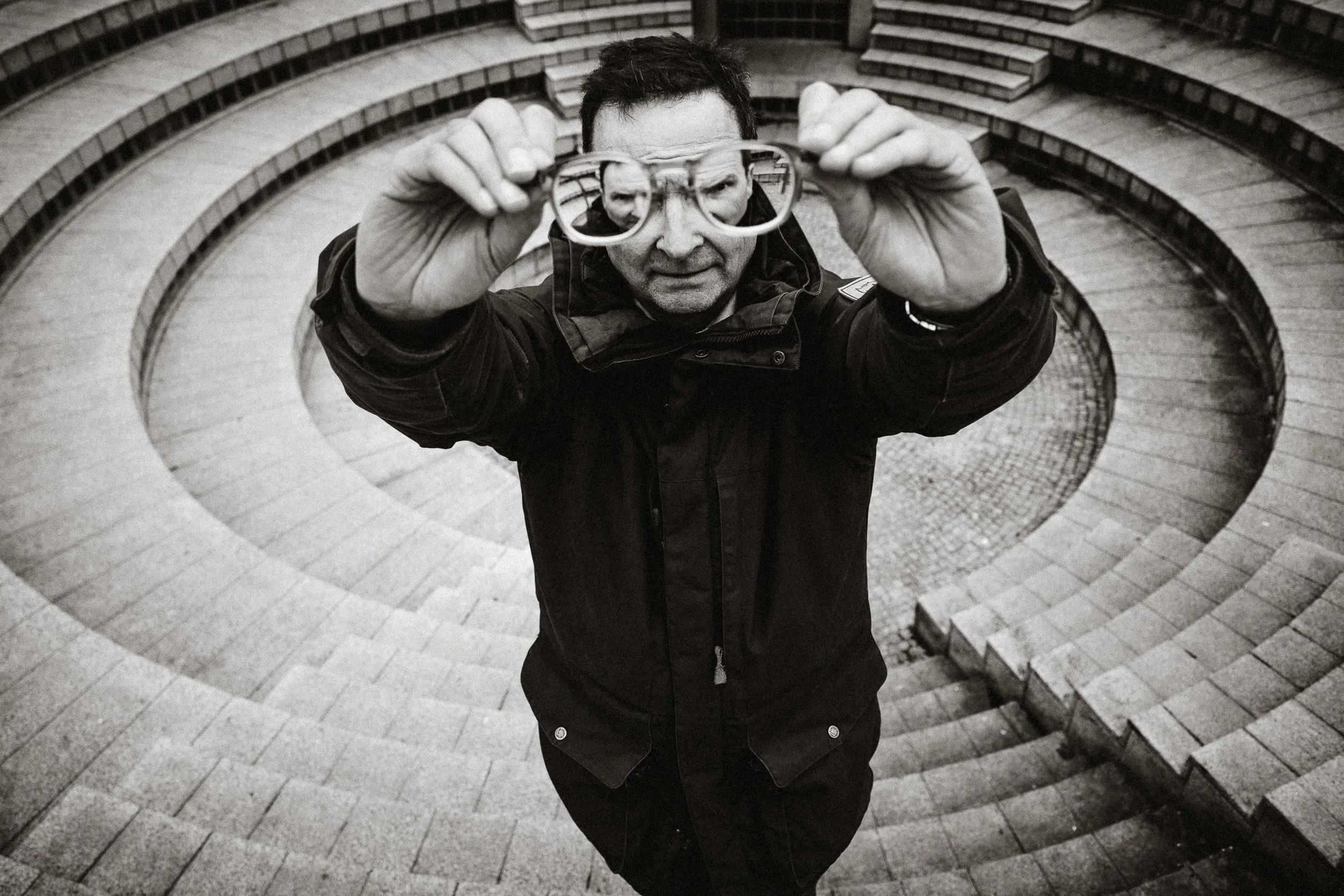 A man wearing a dark jacket holding eyeglasses, standing at the top of spiral stone stairs and looking directly at the camera.