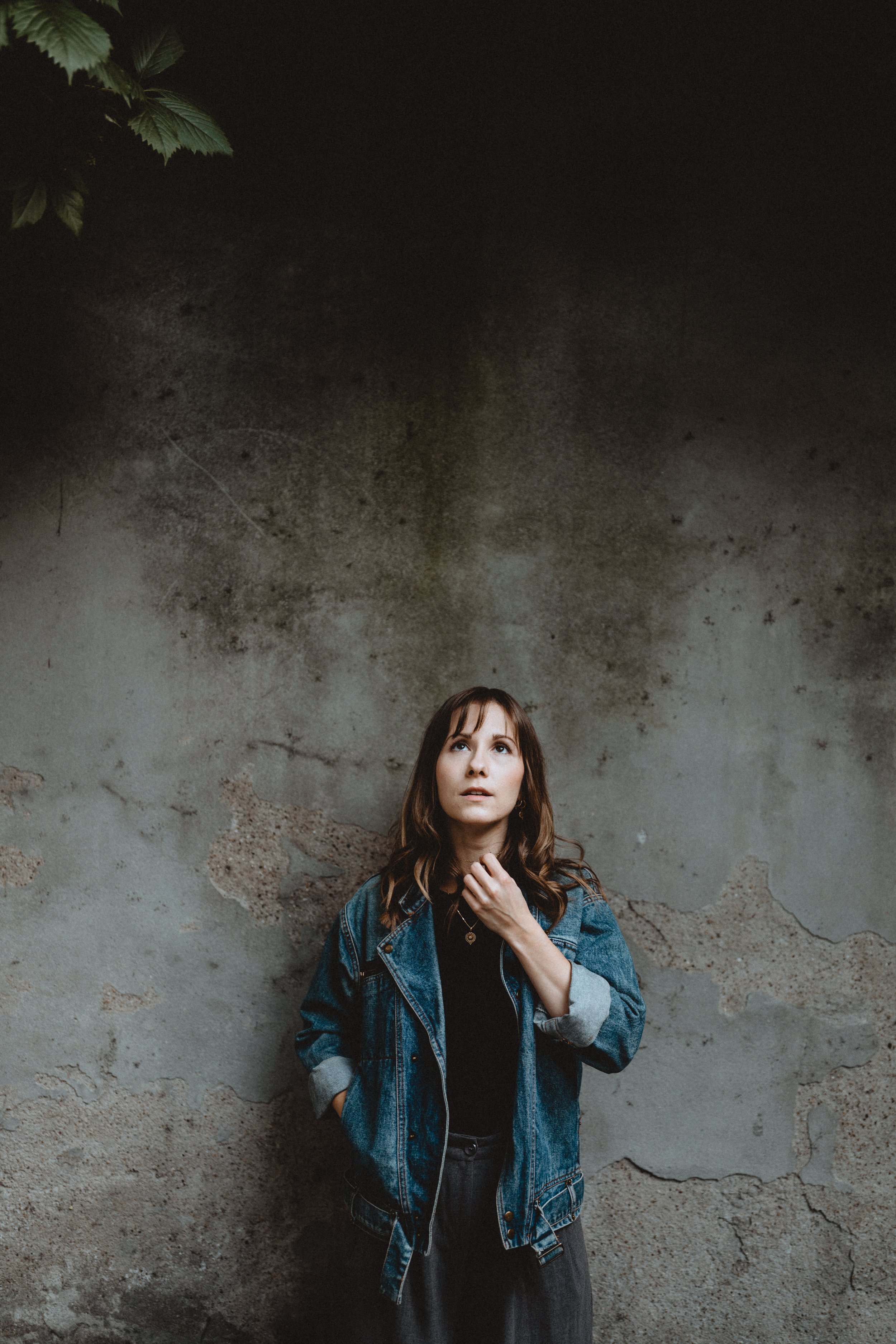 Woman in a denim jacket standing against a weathered concrete wall with a thought expression.