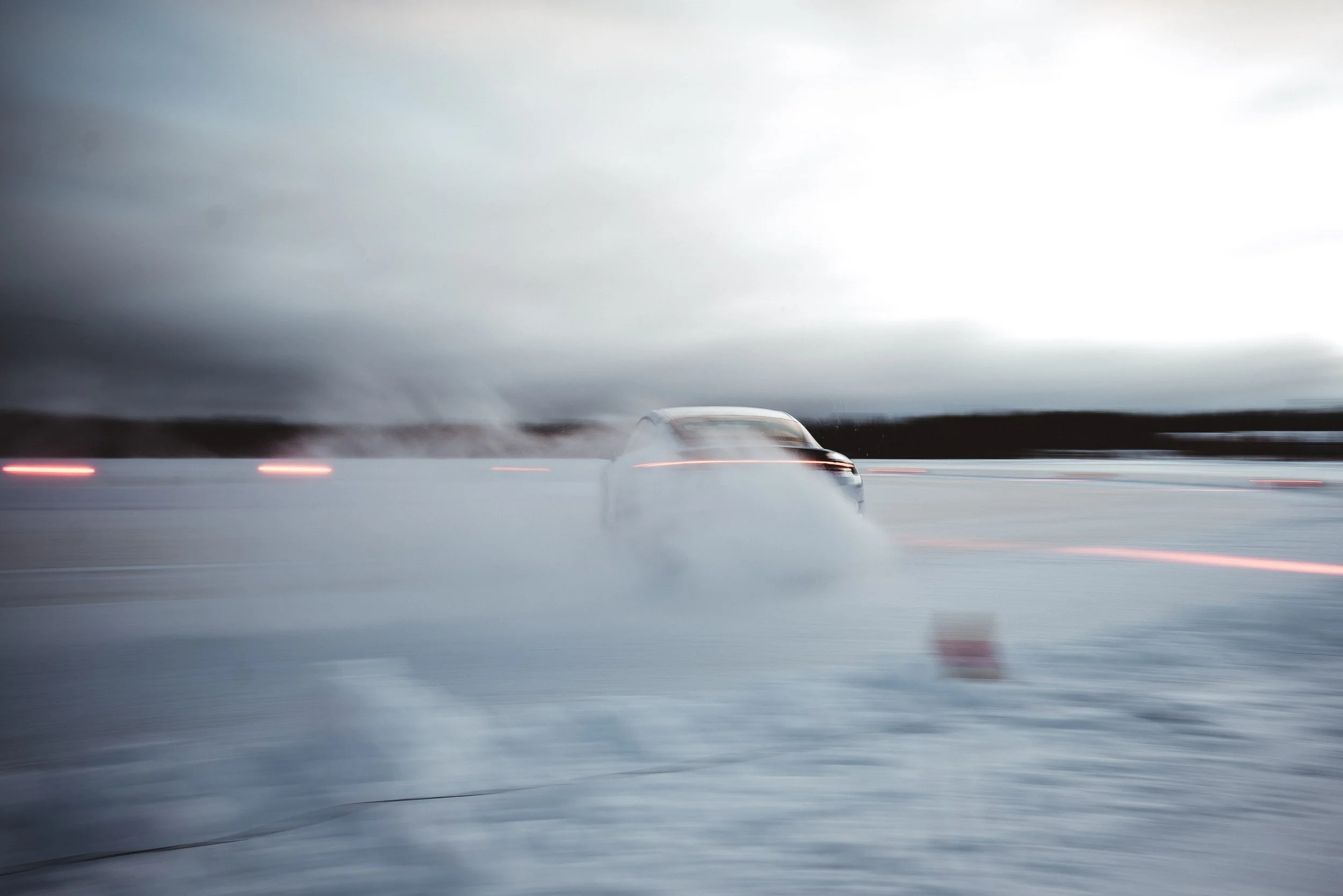 A car driving on a snowy landscape at dusk, kicking up snow and leaving a trail of spray behind it, with blurred background and overcast sky.