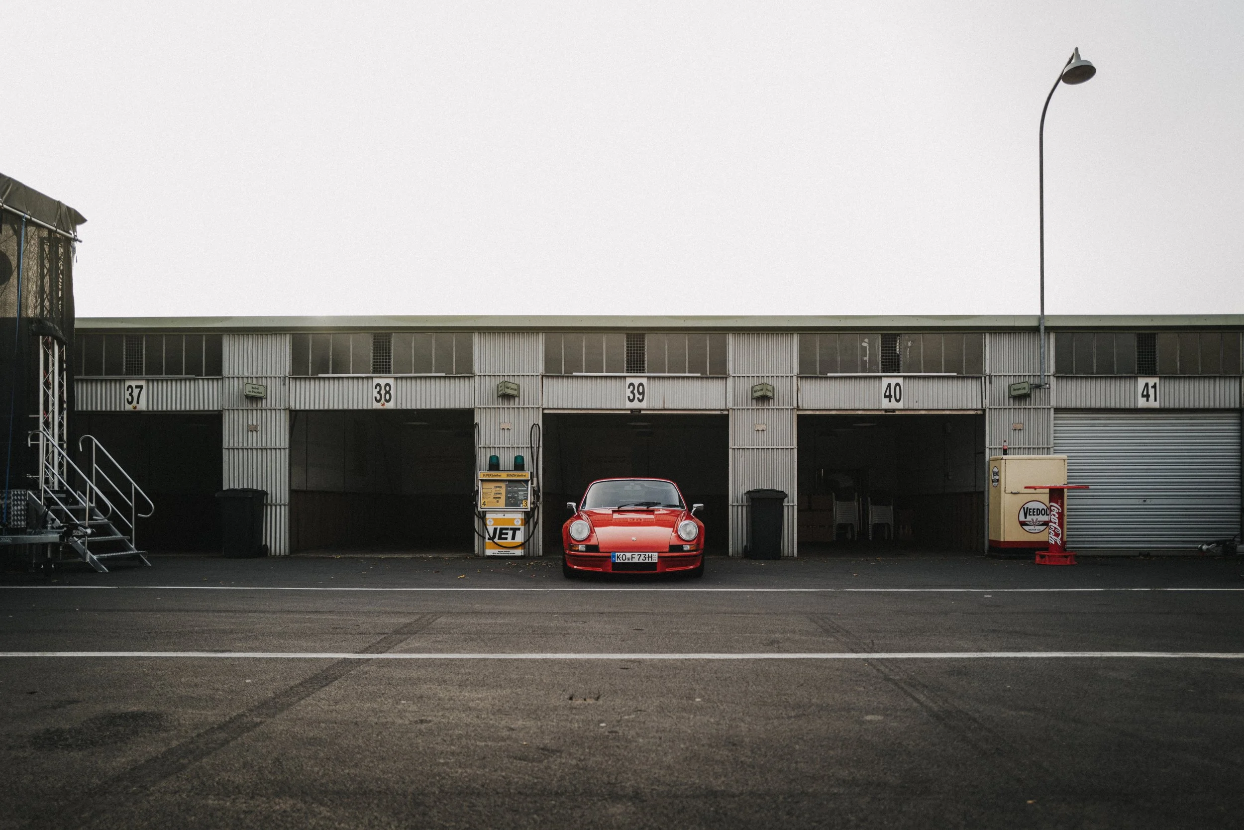 A red sports car parked in front of a garage with open and closed doors numbered 37 to 41, with vintage gas and snack machines nearby.