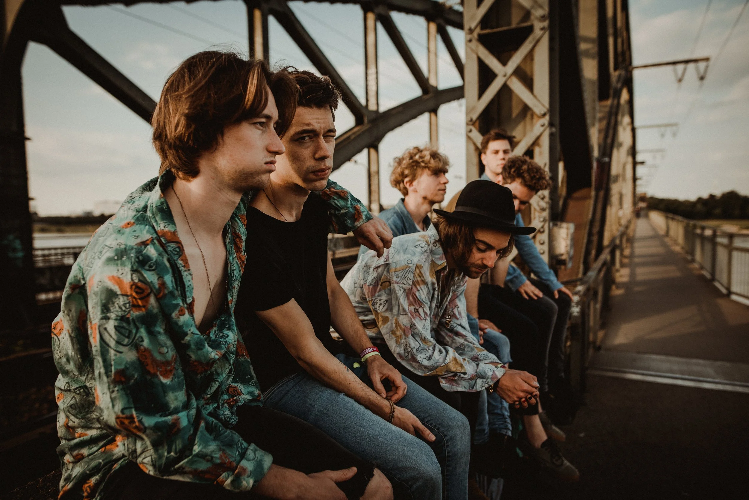 Group of six young men sitting on a metal bench on a bridge, some looking at the camera and others looking down, with a sunset sky in the background.
