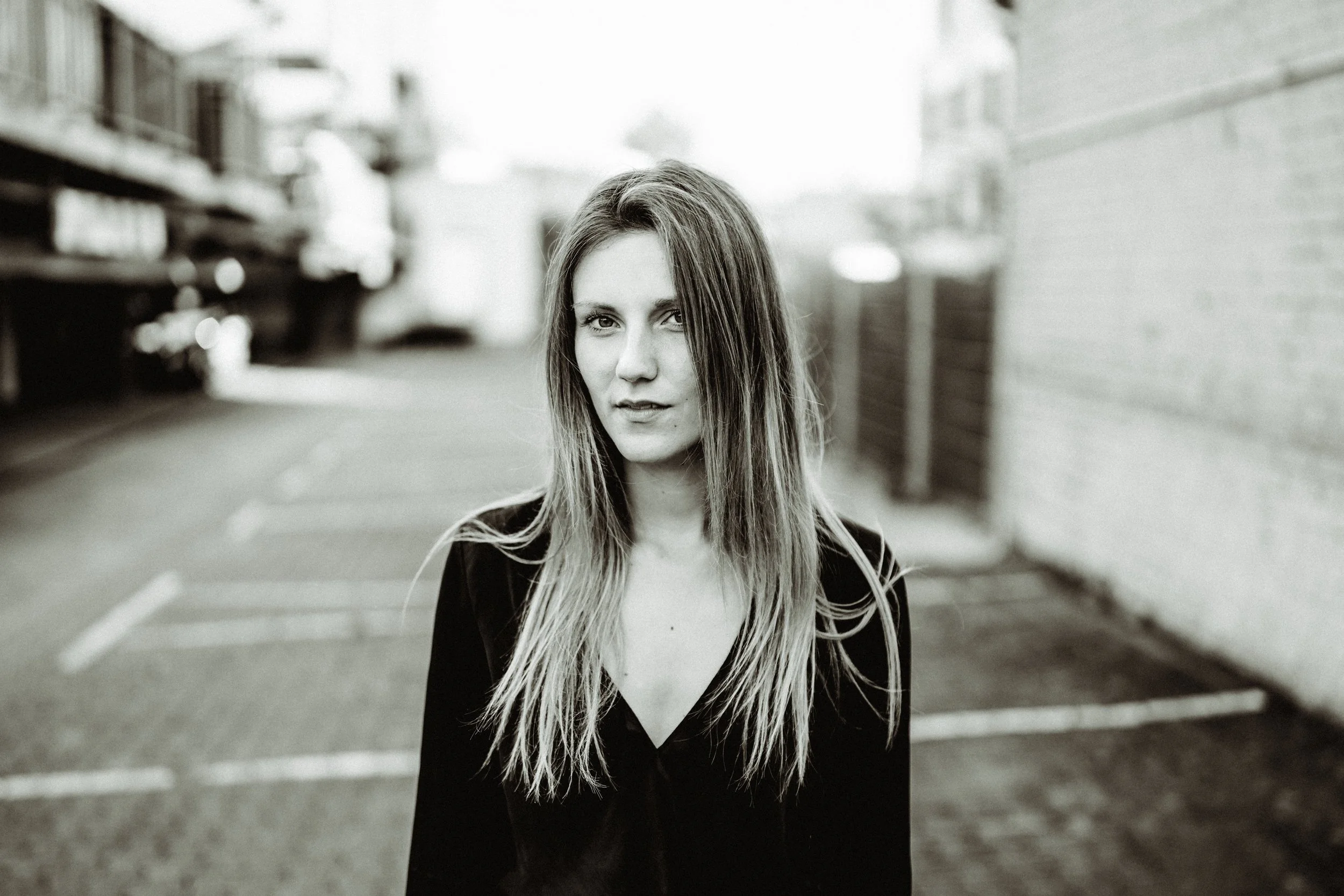 Black and white photo of a woman with long hair standing in an alleyway, looking at the camera with a serious expression.