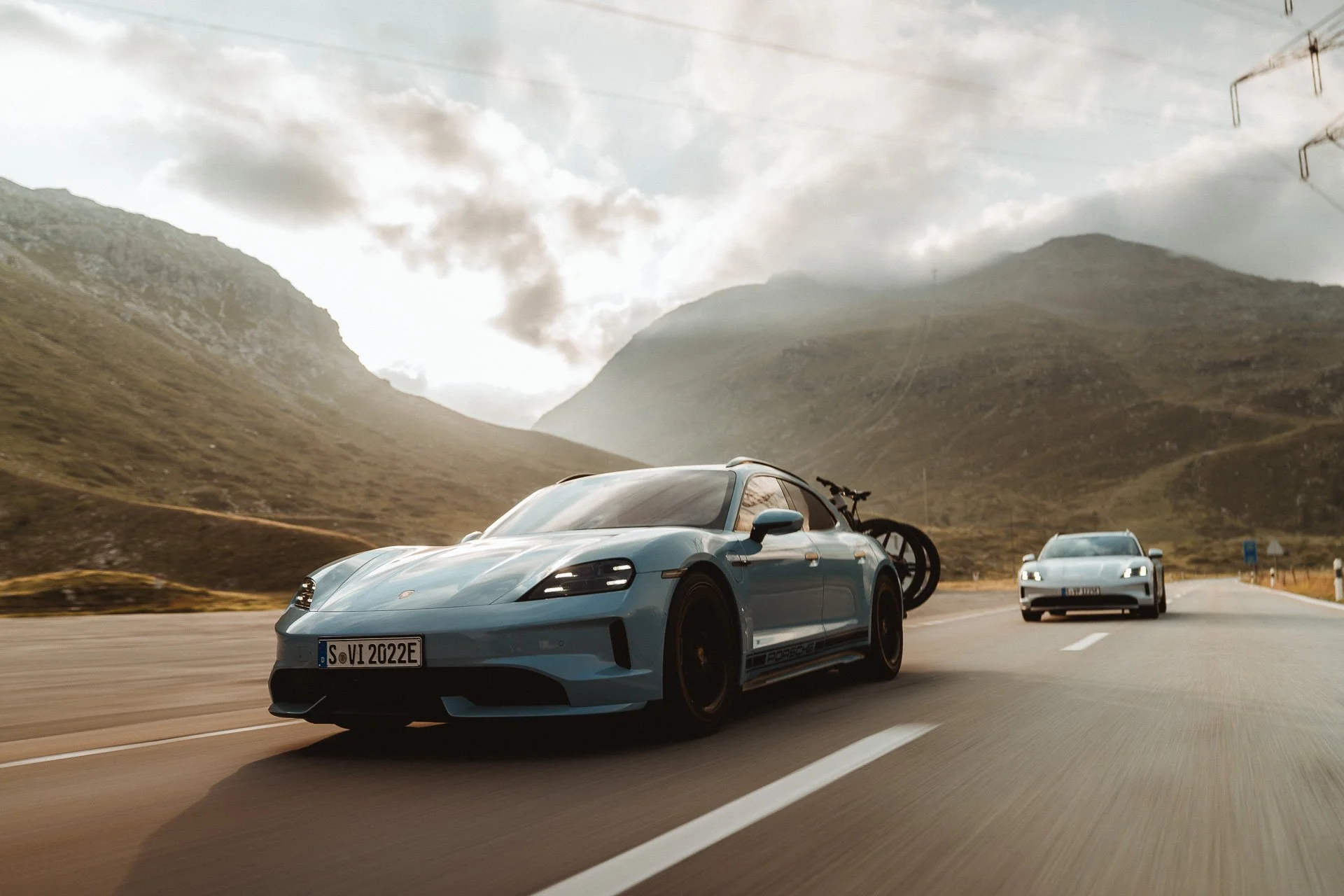 A blue Porsche sports car driving on a mountain highway with a black bicycle mounted on the rear, alongside a silver sports car in the background, with mountains and cloudy sky in the distance.