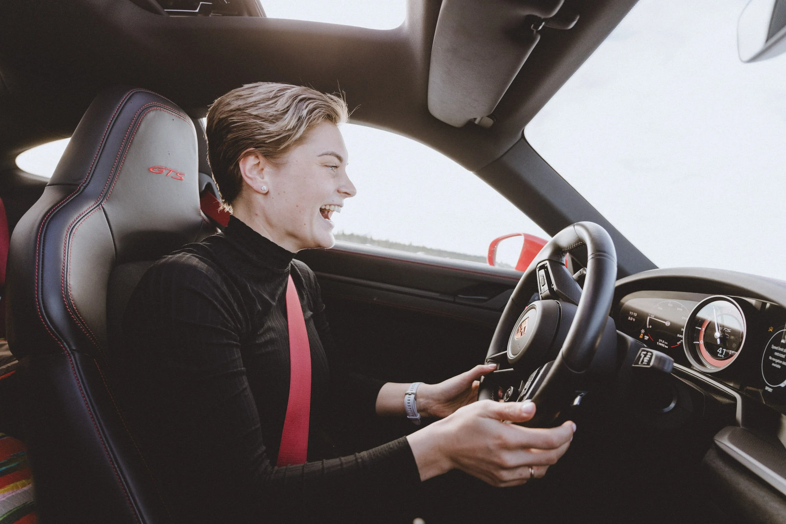 A woman driving a Porsche sports car, smiling and focused on the road, with a grey and red interior.
