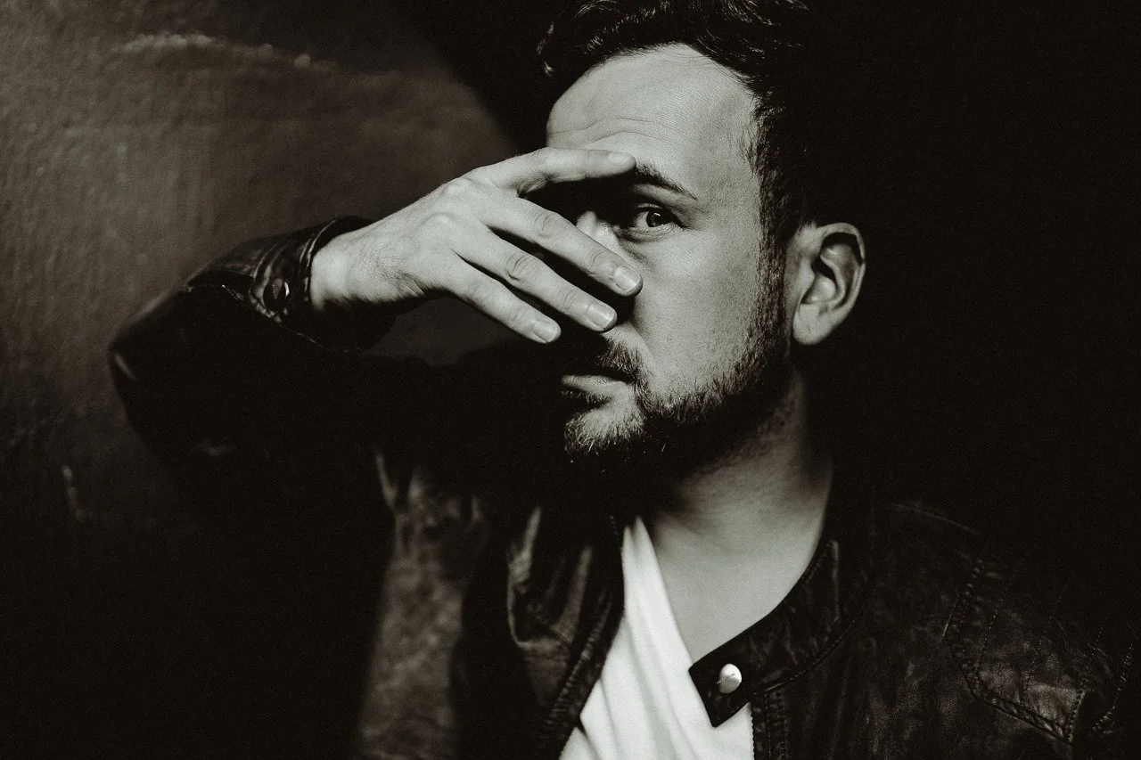 Black and white photo of a man with a beard wearing a leather jacket, covering part of his face with his hand, looking at the camera.