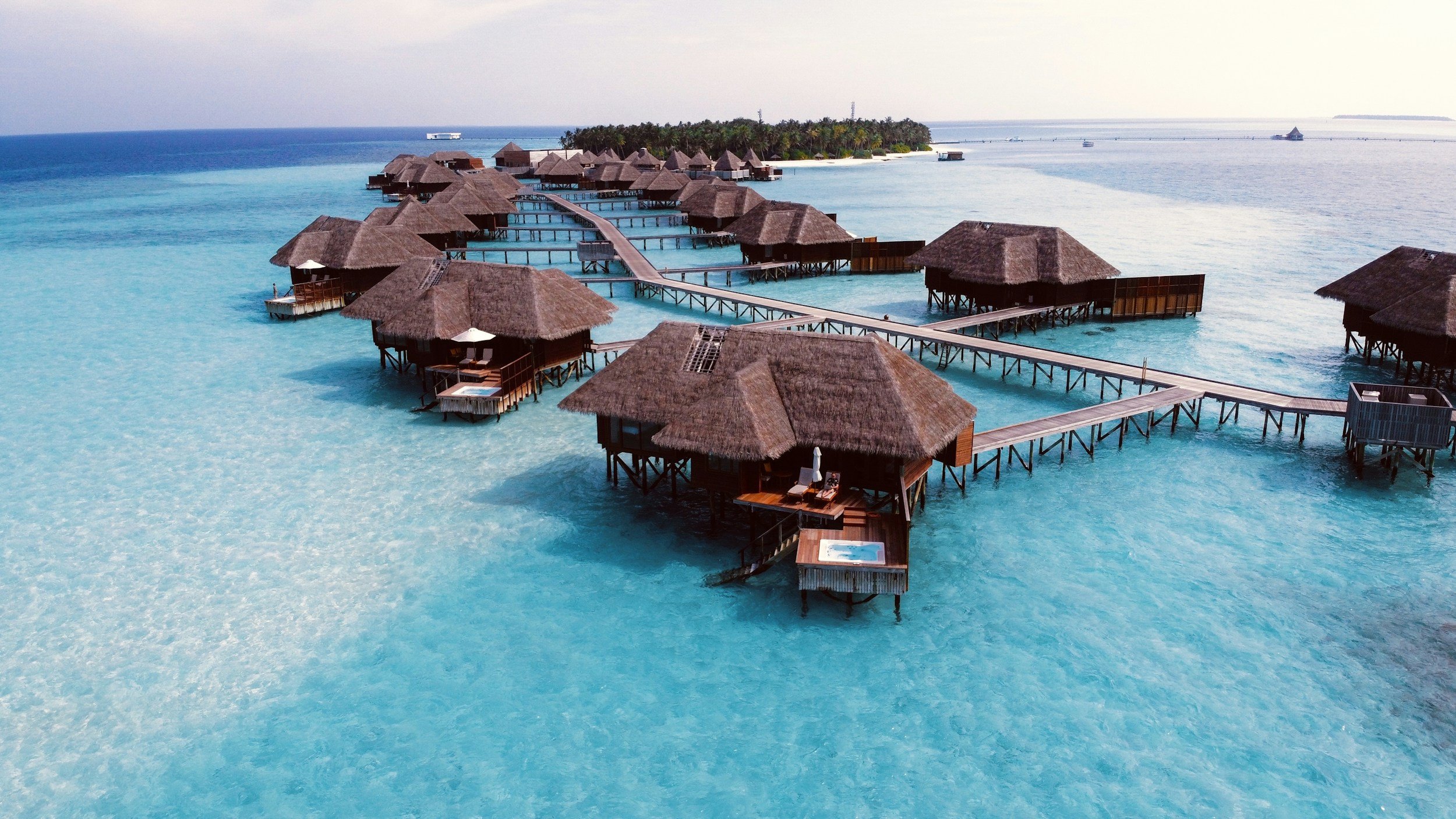 Overwater bungalow resort with thatched roofs, connected by wooden walkways, in crystal-clear blue ocean water with a tropical island in the background.