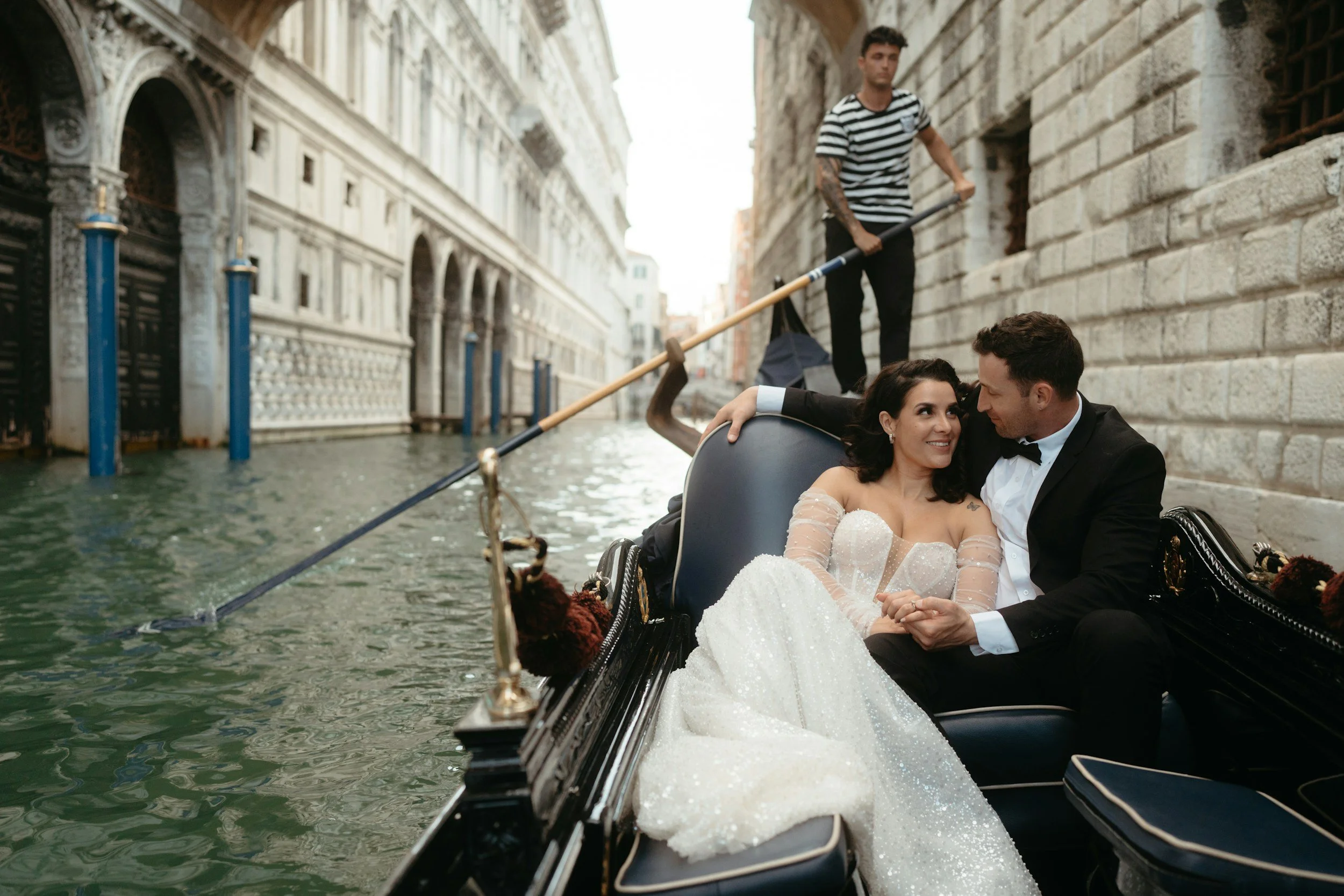 A couple in wedding attire sitting in a gondola on a canal with historic buildings in Venice, Italy, and a gondolier standing at the stern with a pole.