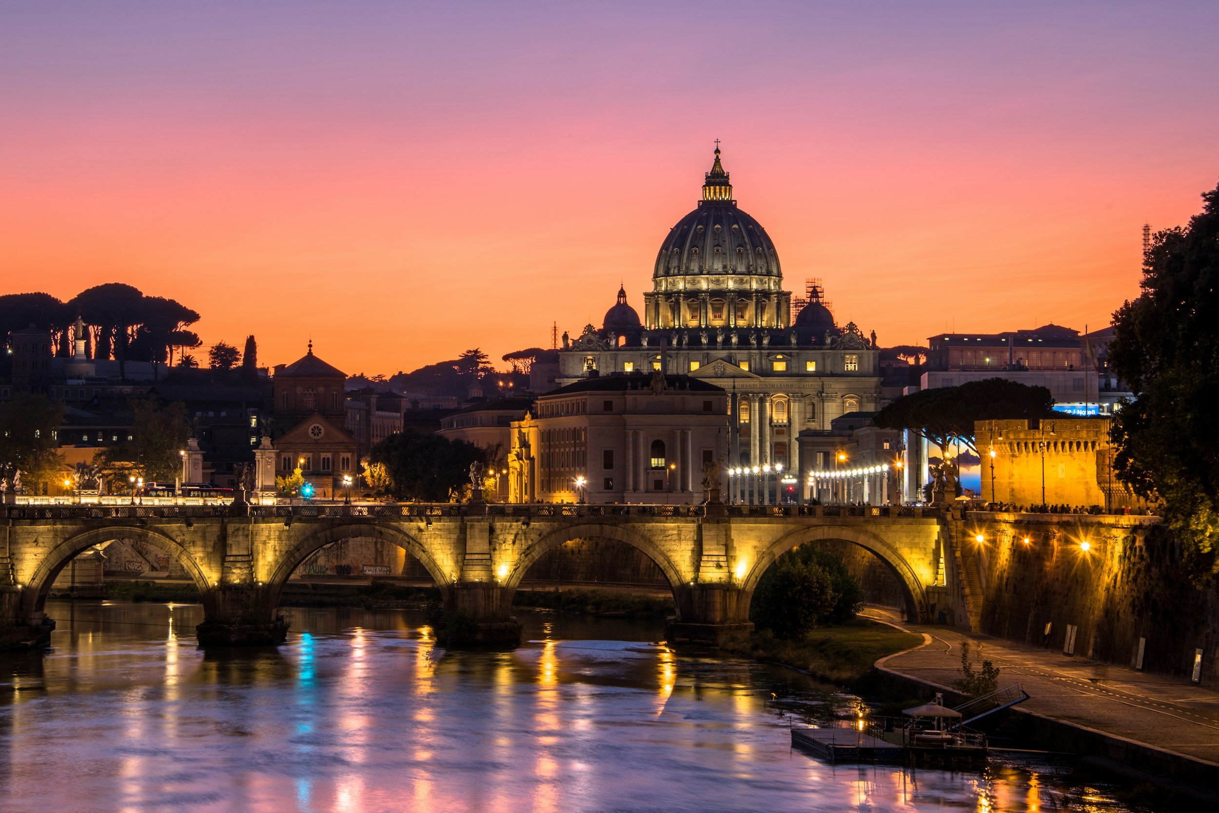 Cityscape of St. Peter's Basilica in Vatican City illuminated at dusk, with reflections on the river below, and a colorful sunset sky in the background.