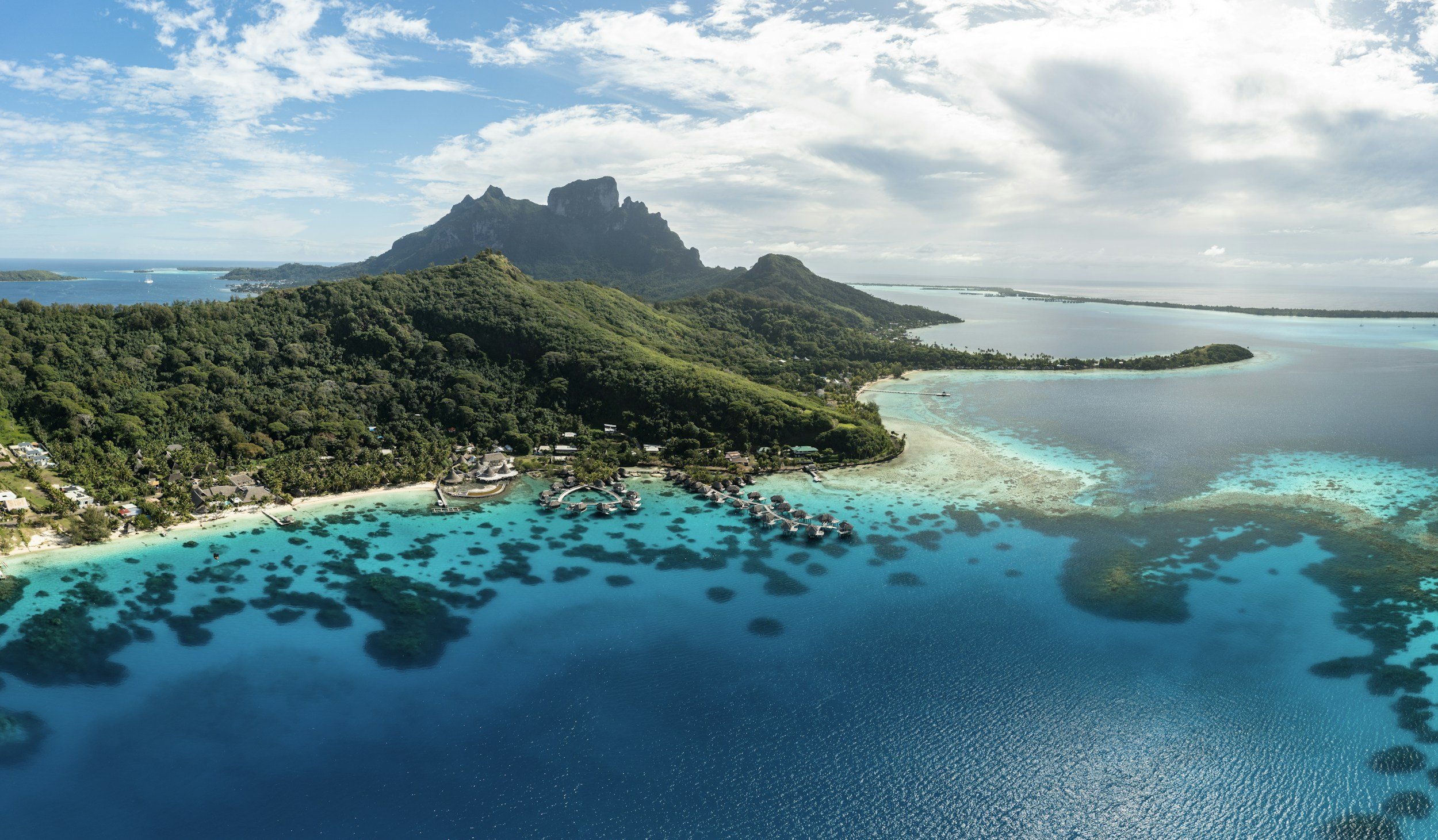 Aerial view of a tropical island with lush green hills, overwater bungalows, and turquoise water with dark patches of coral reef.
