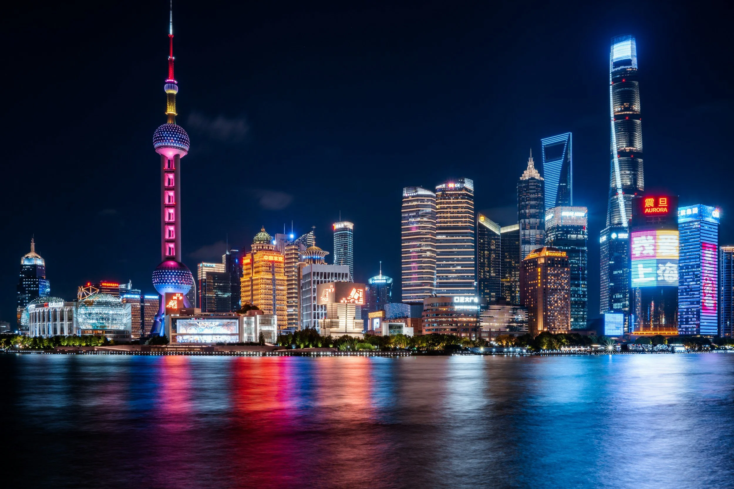 Night view of a illuminated city skyline with skyscrapers reflecting on water, including the Oriental Pearl Tower in Shanghai.