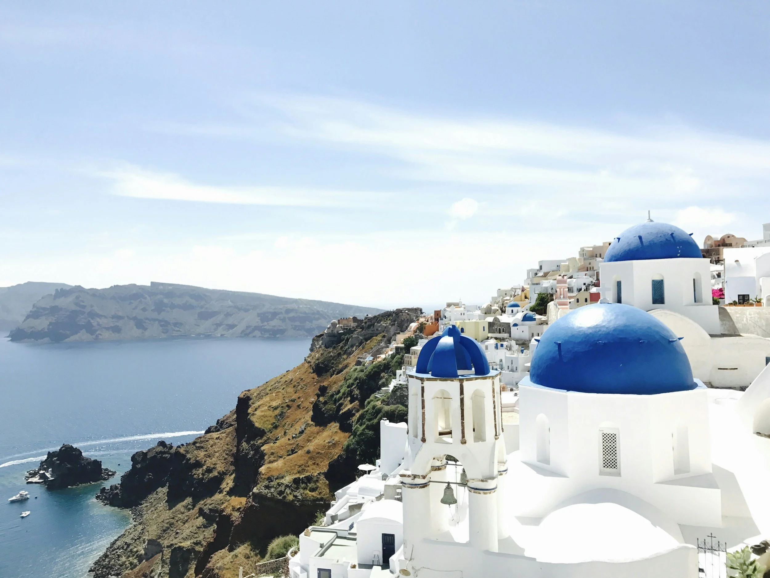 White buildings with blue domes on a cliffside overlooking the ocean, against a partly cloudy sky in Santorini, Greece.