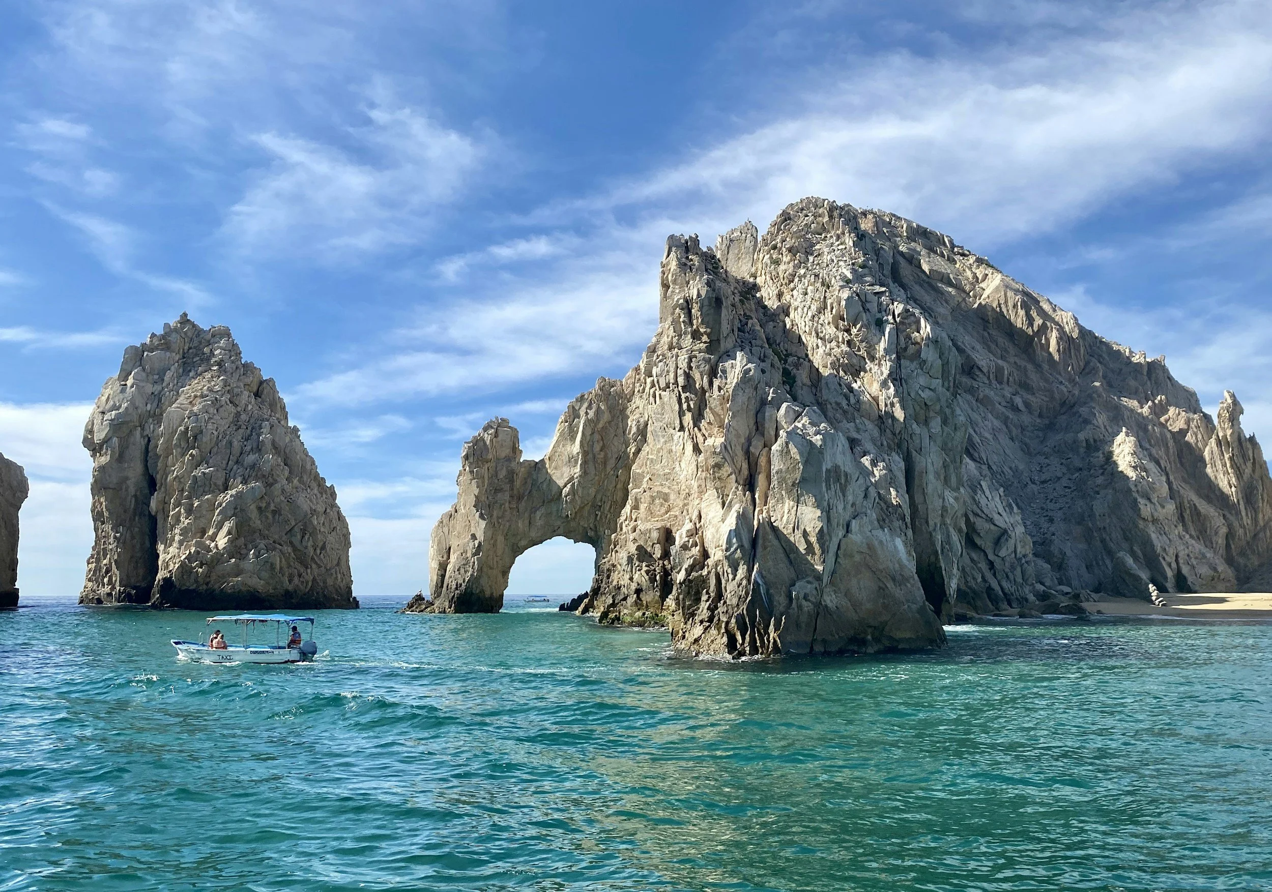 A coastal scene with large rock formations, including an arch, under a blue sky with wispy clouds. A boat with two people is floating on clear turquoise water near the rocks.