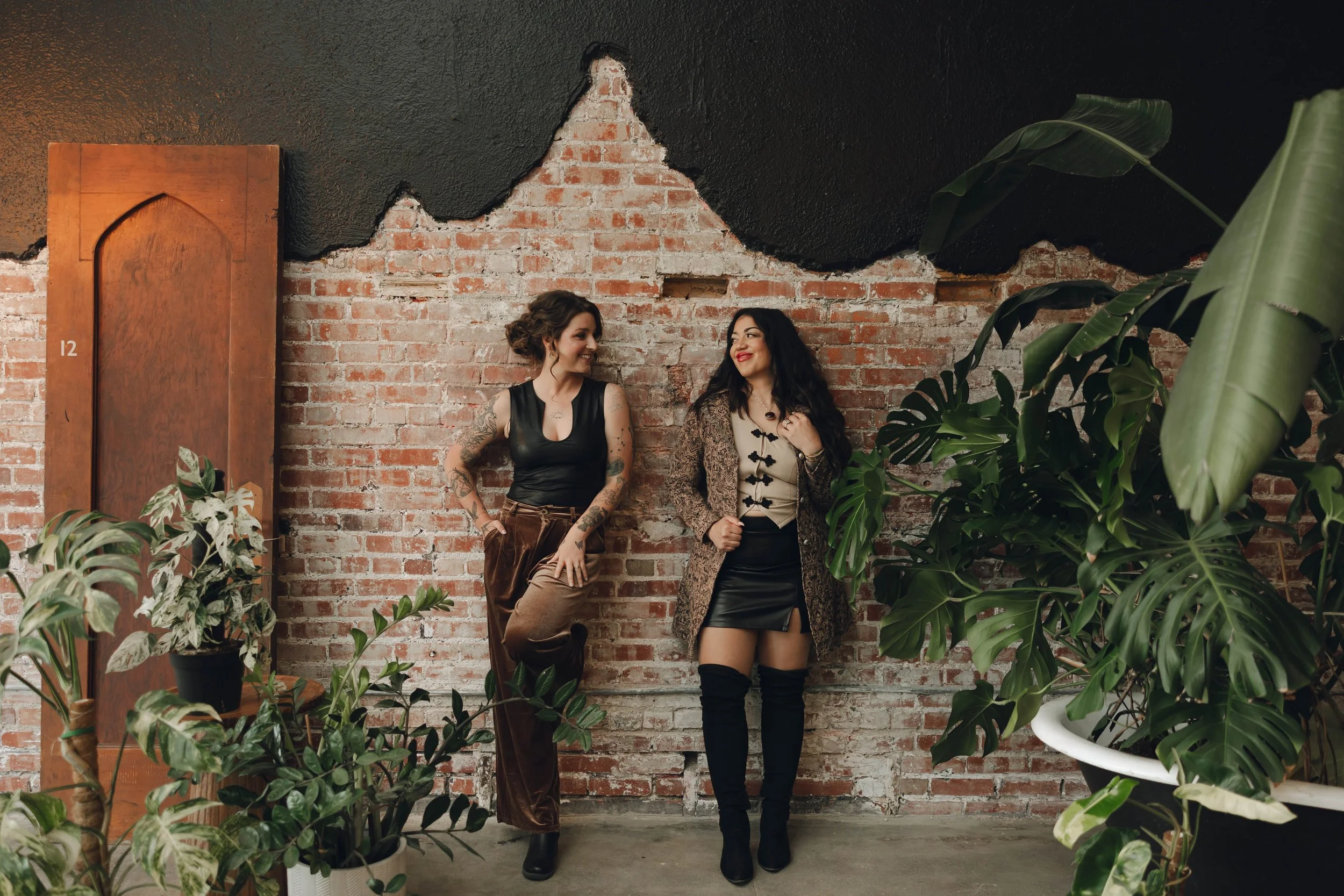 Two women standing and smiling in front of a brick wall with black paint and large green plants around them.