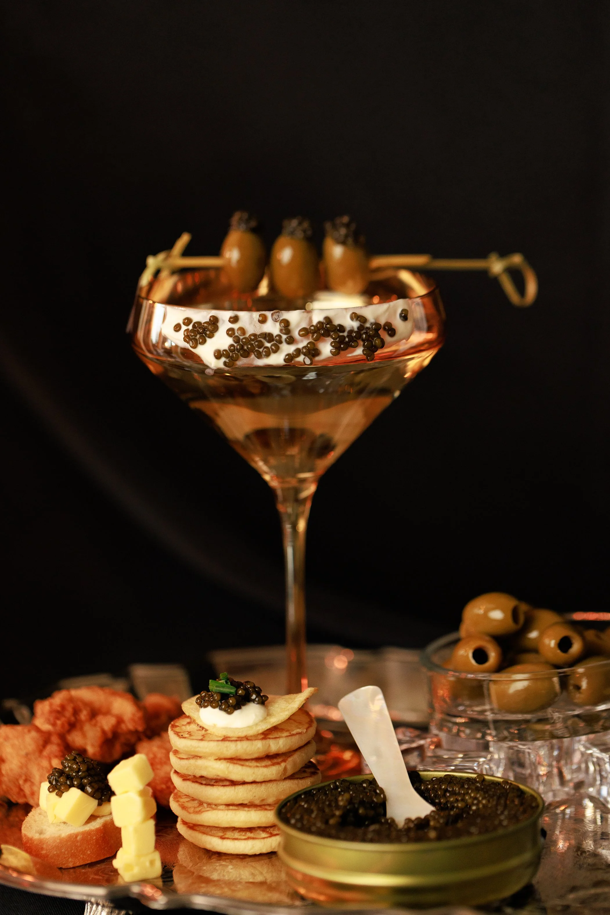 A fancy cocktail glass with a drink garnished with black caviar, and appetizers including stacked pancakes topped with caviar, cheese cubes, and other finger foods, with bowls of green olives and caviar on a tray, all set against a dark background.