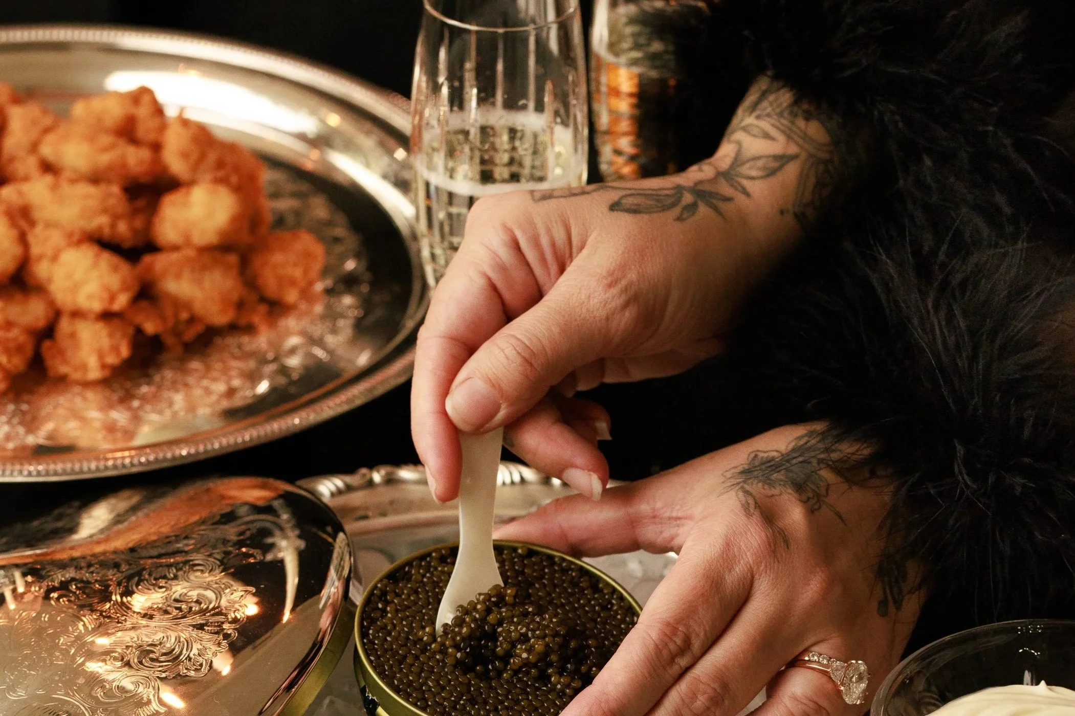 A person with tattoos on their hands and a ring on their finger is scooping caviar from a small container with a white spoon, at a table with various dishes and drinks, including fried food and a glass of clear beverage.